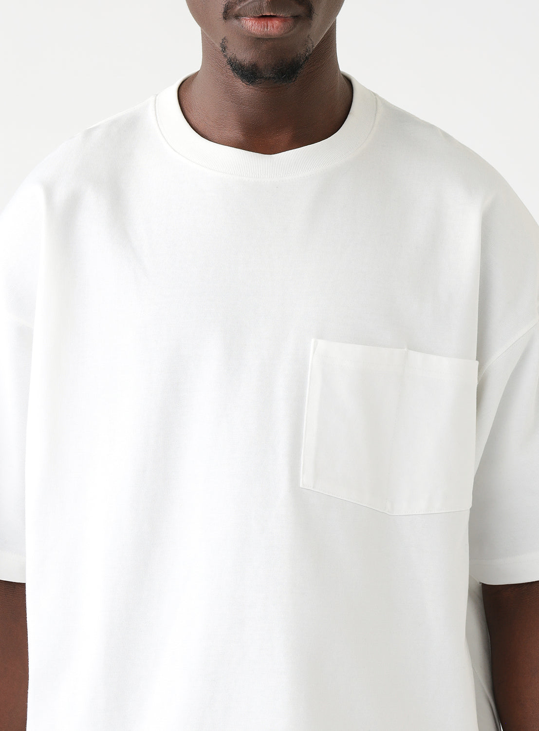 Man wearing a white oversized T-shirt with a chest pocket and gray cargo pants, standing against a plain white background. The T-shirt features a relaxed fit with dropped shoulders and a crew neckline.
