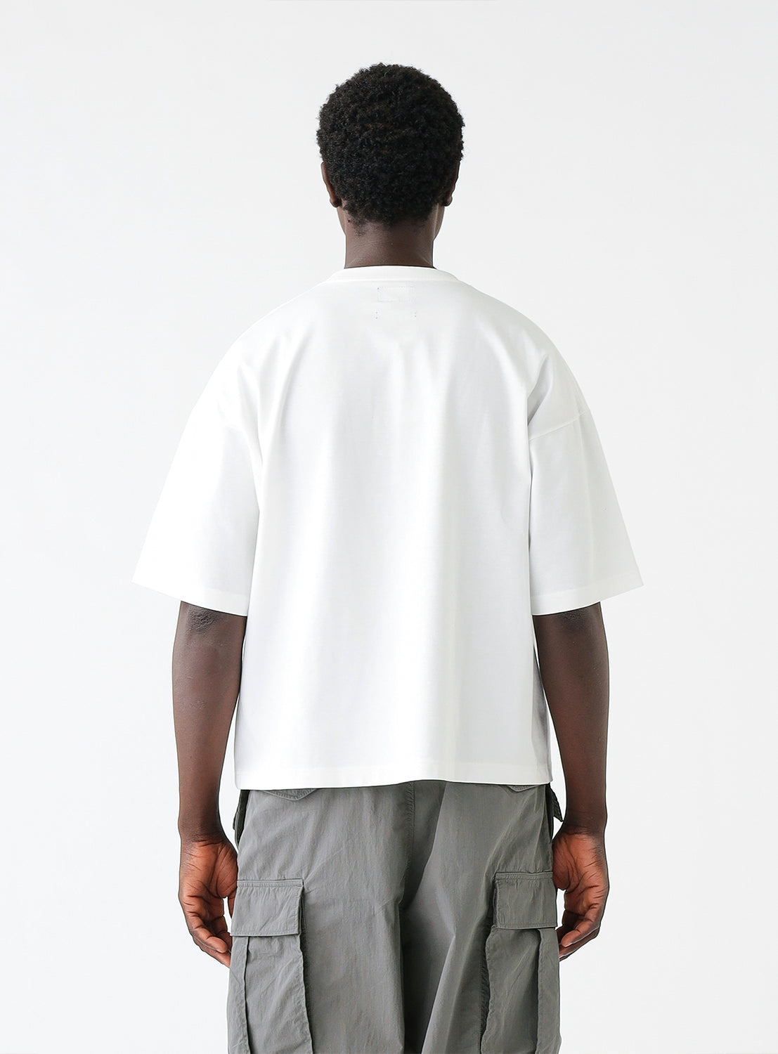 Man wearing a white oversized T-shirt with a chest pocket and gray cargo pants, standing against a plain white background. The T-shirt features a relaxed fit with dropped shoulders and a crew neckline.