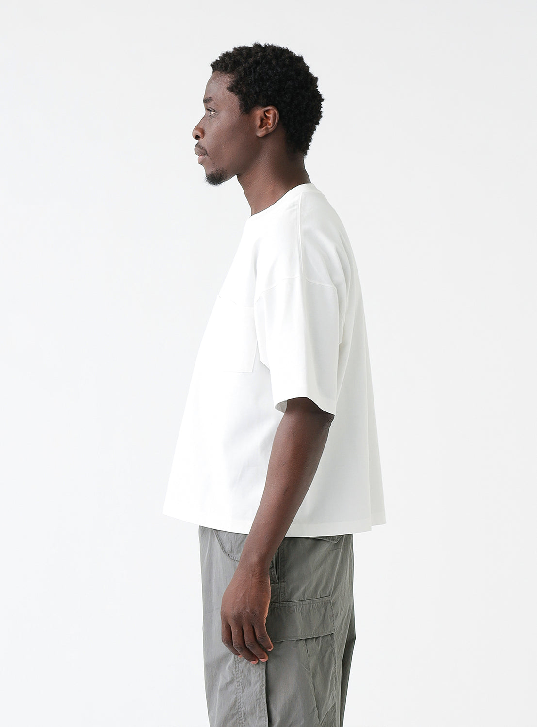 Man wearing a white oversized T-shirt with a chest pocket and gray cargo pants, standing against a plain white background. The T-shirt features a relaxed fit with dropped shoulders and a crew neckline.