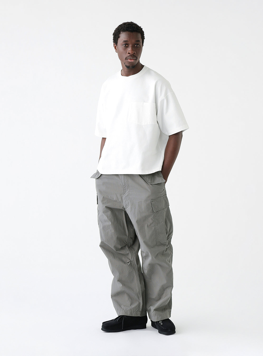 Man wearing a white oversized T-shirt with a chest pocket and gray cargo pants, standing against a plain white background. The T-shirt features a relaxed fit with dropped shoulders and a crew neckline.