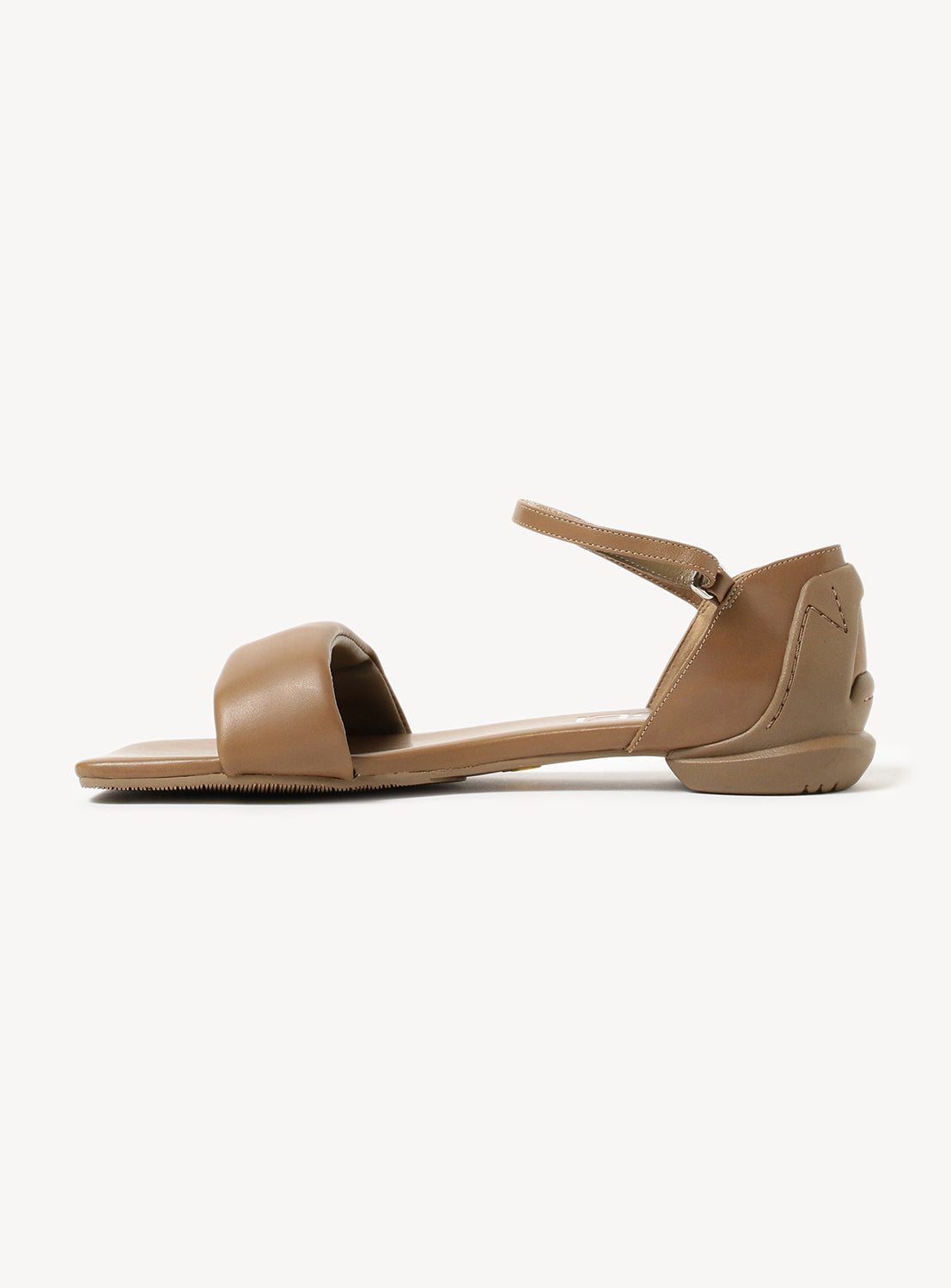A tan flat sandal, shown from the side, with a visible ankle strap and toe strap.