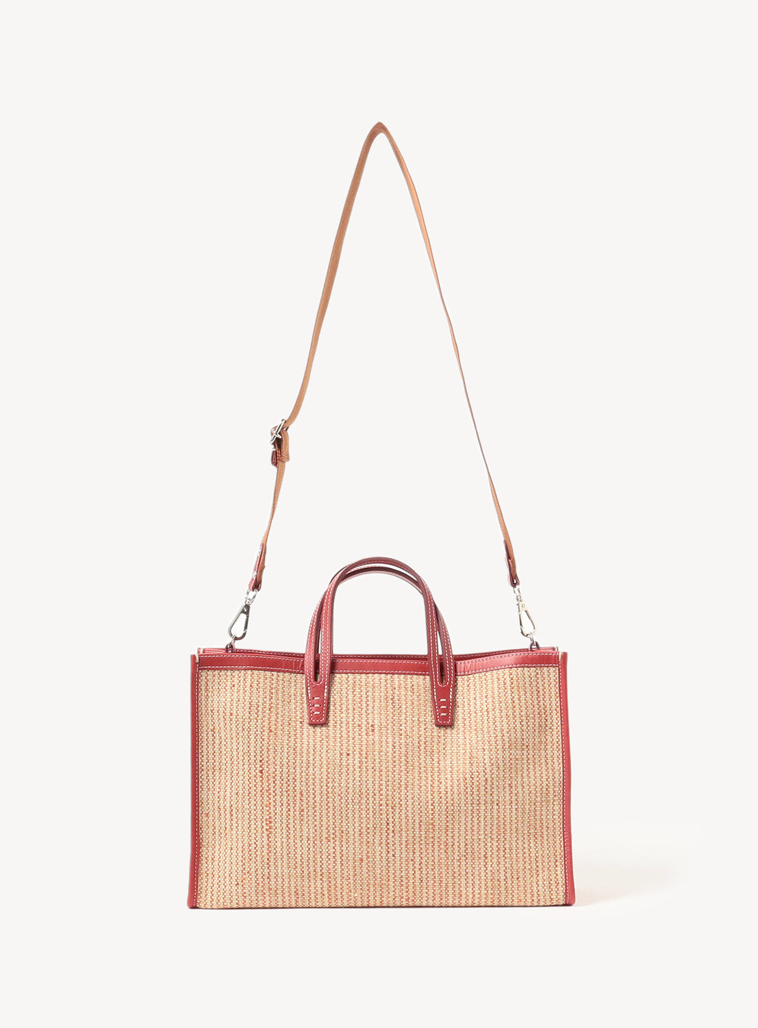A large tote bag made of woven raffia with red leather handles, a long shoulder strap and a trim.