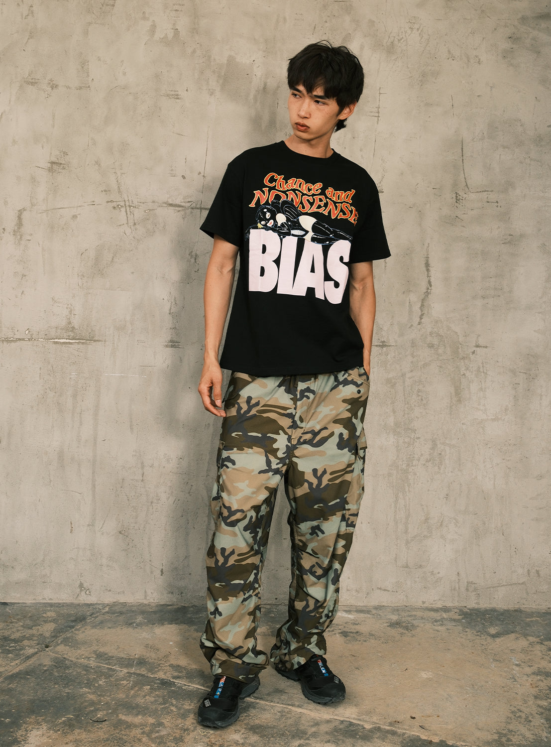 A person stands against a textured concrete wall, wearing the BIAS DOGS 'CRAP' Graphic Tee paired with camo cargo pants and black sneakers—a bold look inspired by Kei Tanaka’s streetwear style.