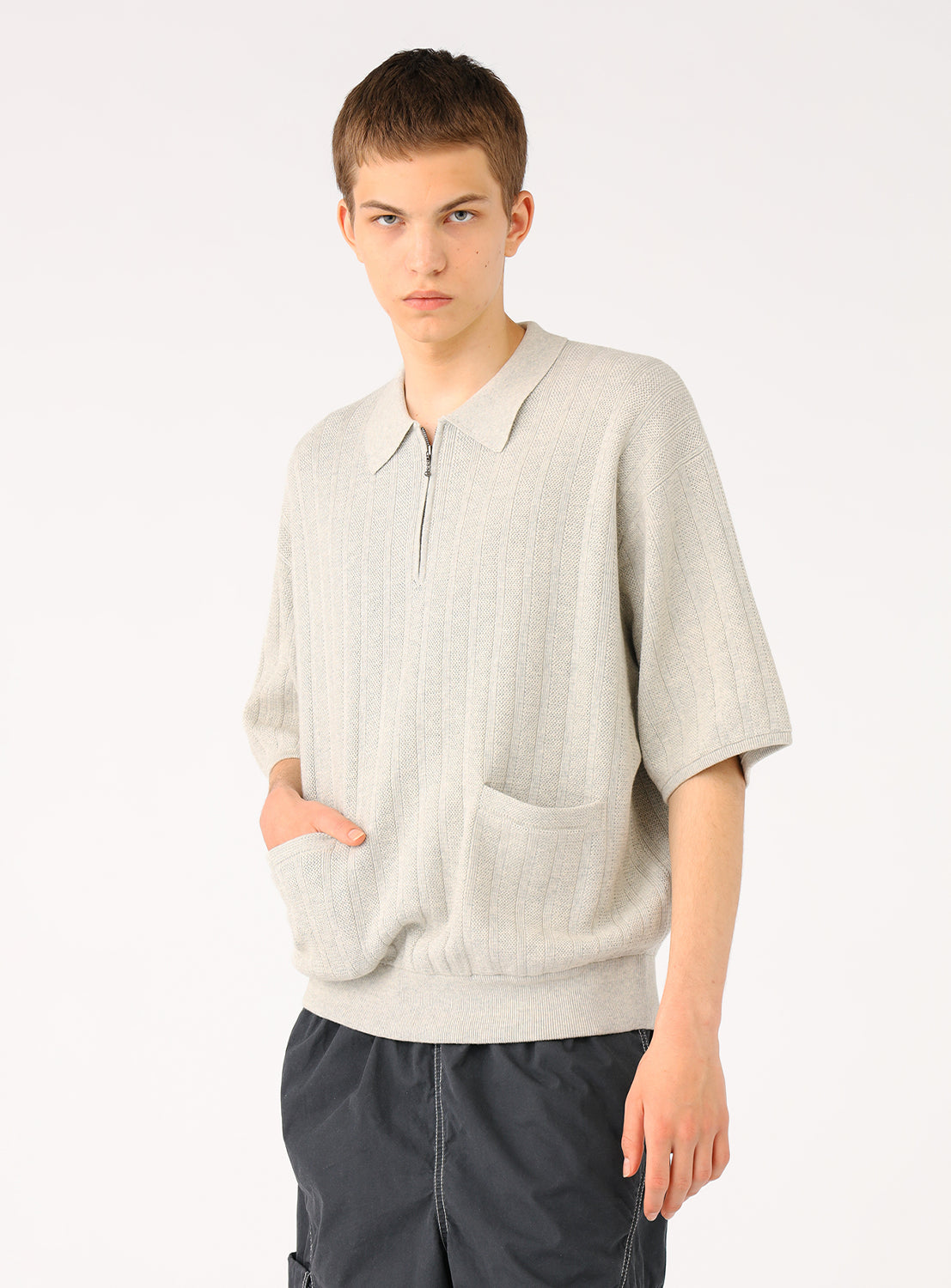 Male model in light grey knit polo and dark shorts, hands in pockets, looking at viewer.