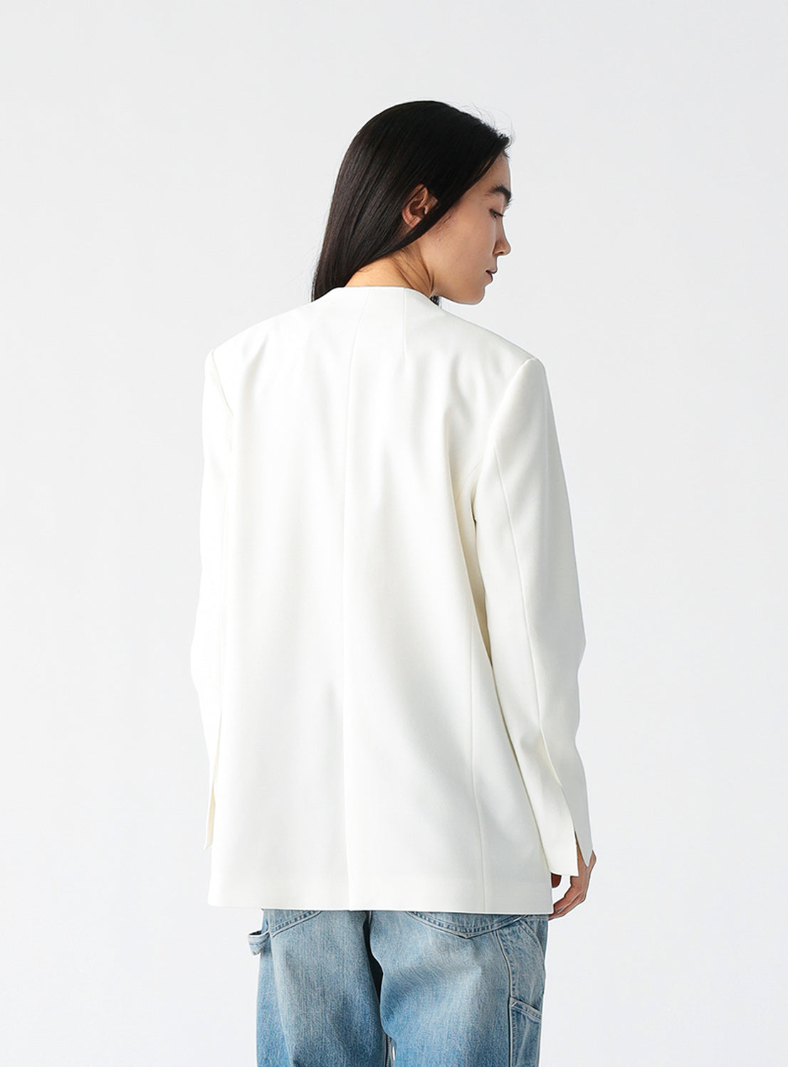 A person wearing a minimalist white blazer with a deep V-neckline and structured shoulders, styled over a sheer beige top and paired with light blue patchwork jeans, standing against a plain white background.