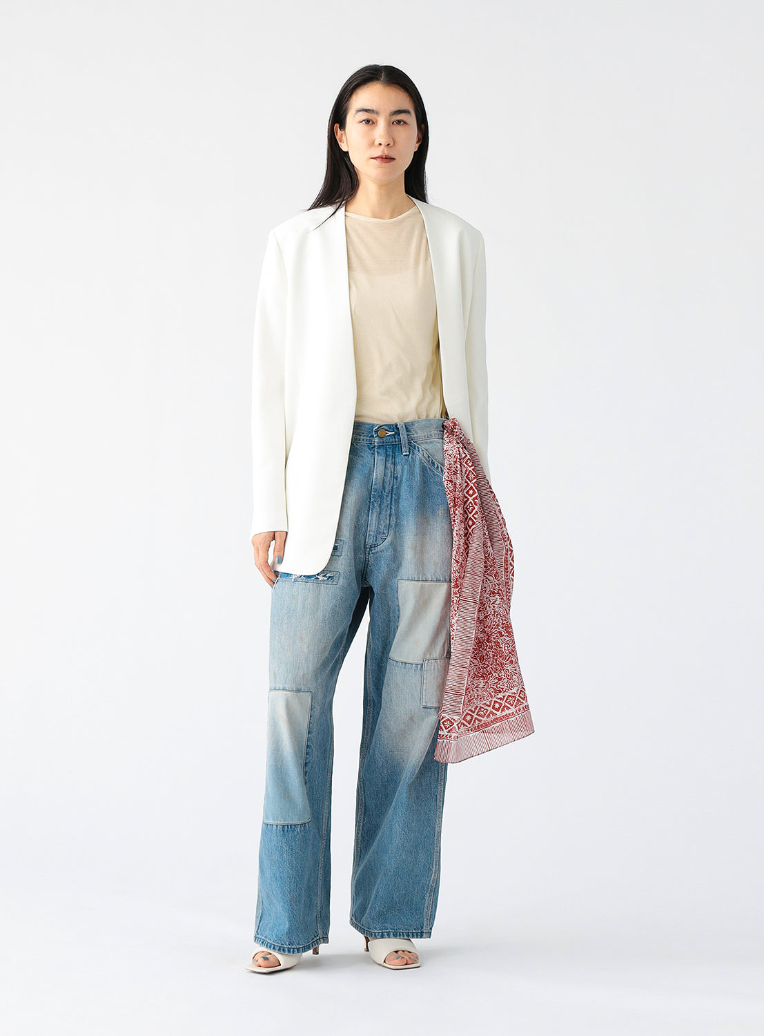 A person wearing a minimalist white blazer with a deep V-neckline and structured shoulders, styled over a sheer beige top and paired with light blue patchwork jeans, standing against a plain white background.