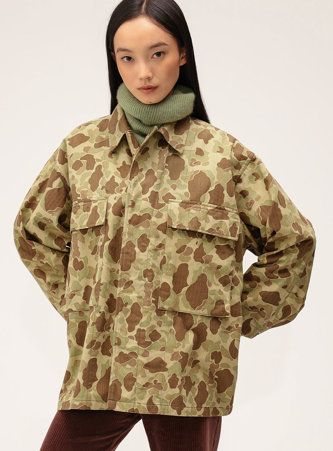 A woman stands with her hands on her hips, wearing the Buzz Rickson's x BEAMS Boy Herringbone Camo Jacket SP over a green turtleneck sweater against a plain background.