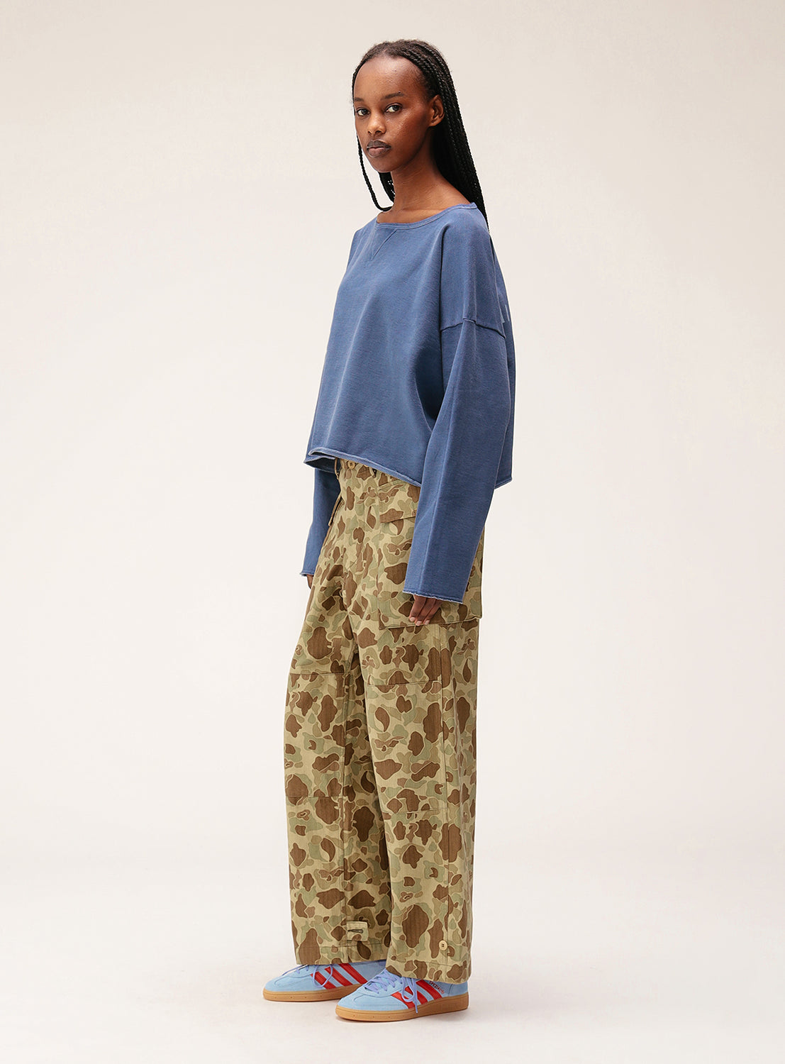 A person with long braided hair and a neutral expression stands against a plain background in a loose blue top, light blue sneakers with red accents, and Buzz Rickson's x BEAMS Boy Herringbone Camo Pants. Made in Japan.
