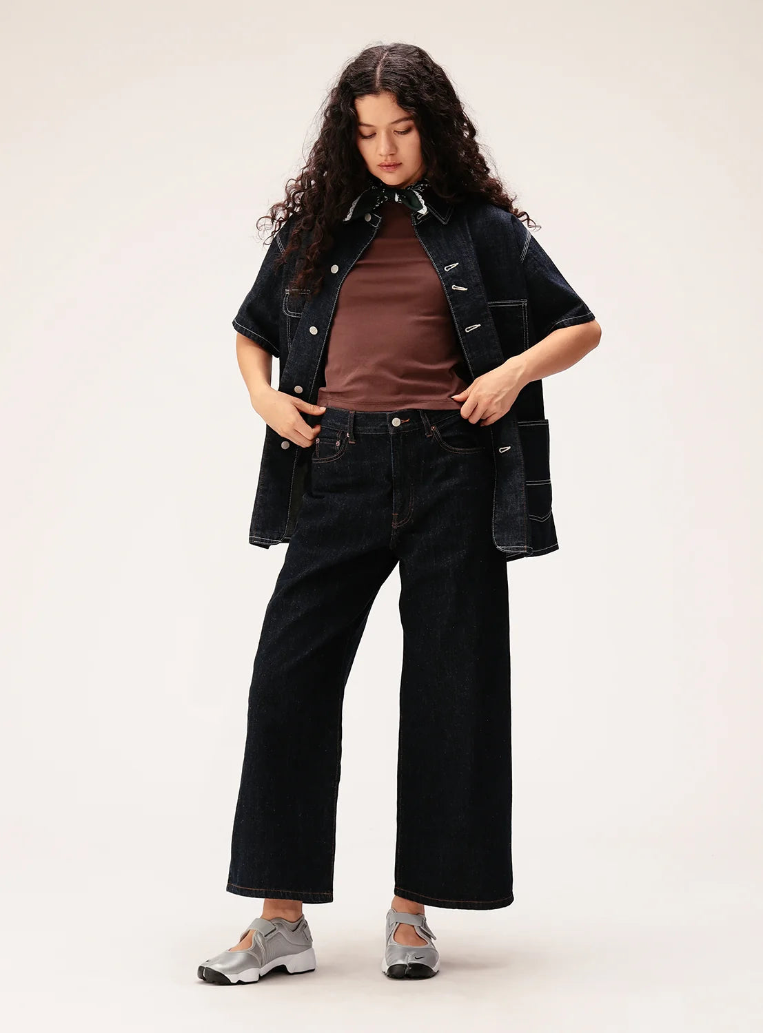 A person with long curly hair wears a brown shirt, a dark denim jacket, BEAMS BOY Straight Fit Wide Denim Pants, and silver shoes, standing against a plain background and looking down with hands in pockets.