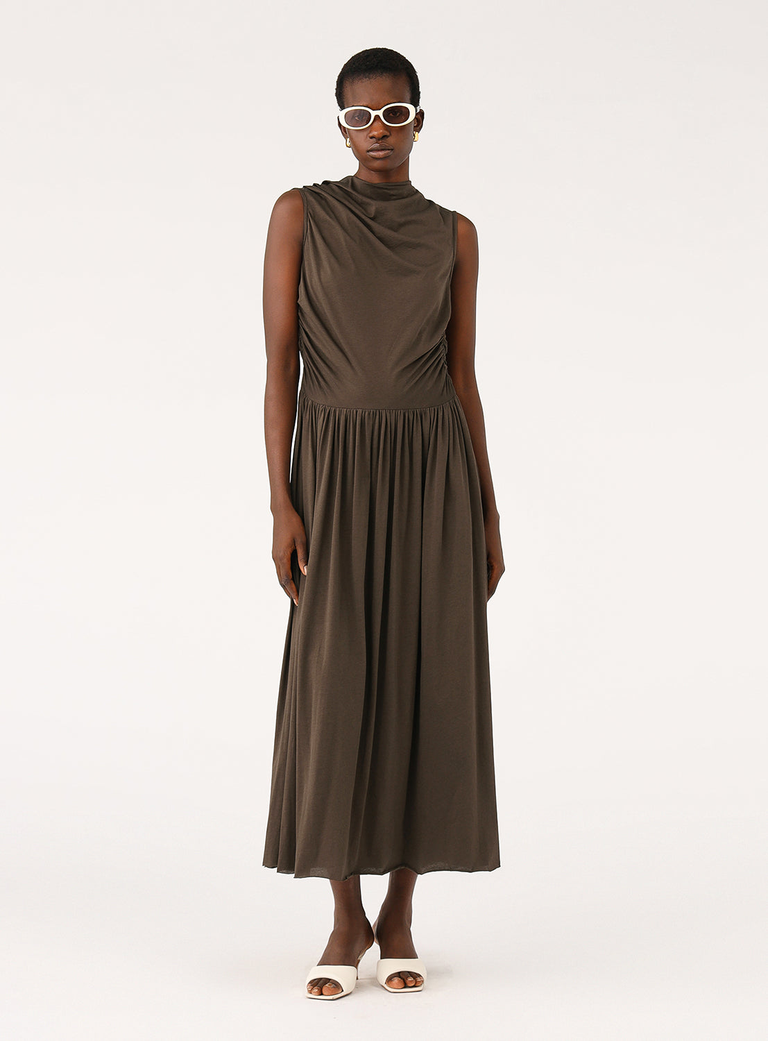 Model wearing a sleeveless brown dress, featuring a high draped neckline and a cinched waist.