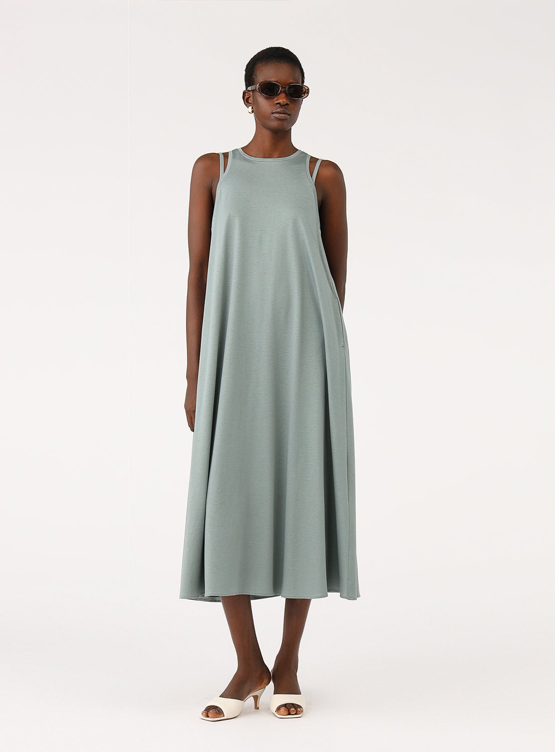 Full body shot of a model wearing a simple sage green A-line midi dress, styled with sandals and sunglasses.