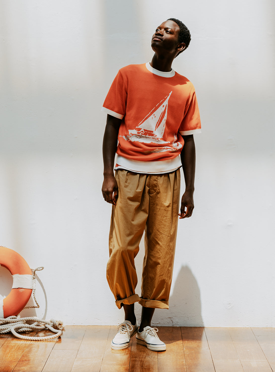 A model wearing an orange crew neck knit tee with a sailboat graphic on the front and brown pants.