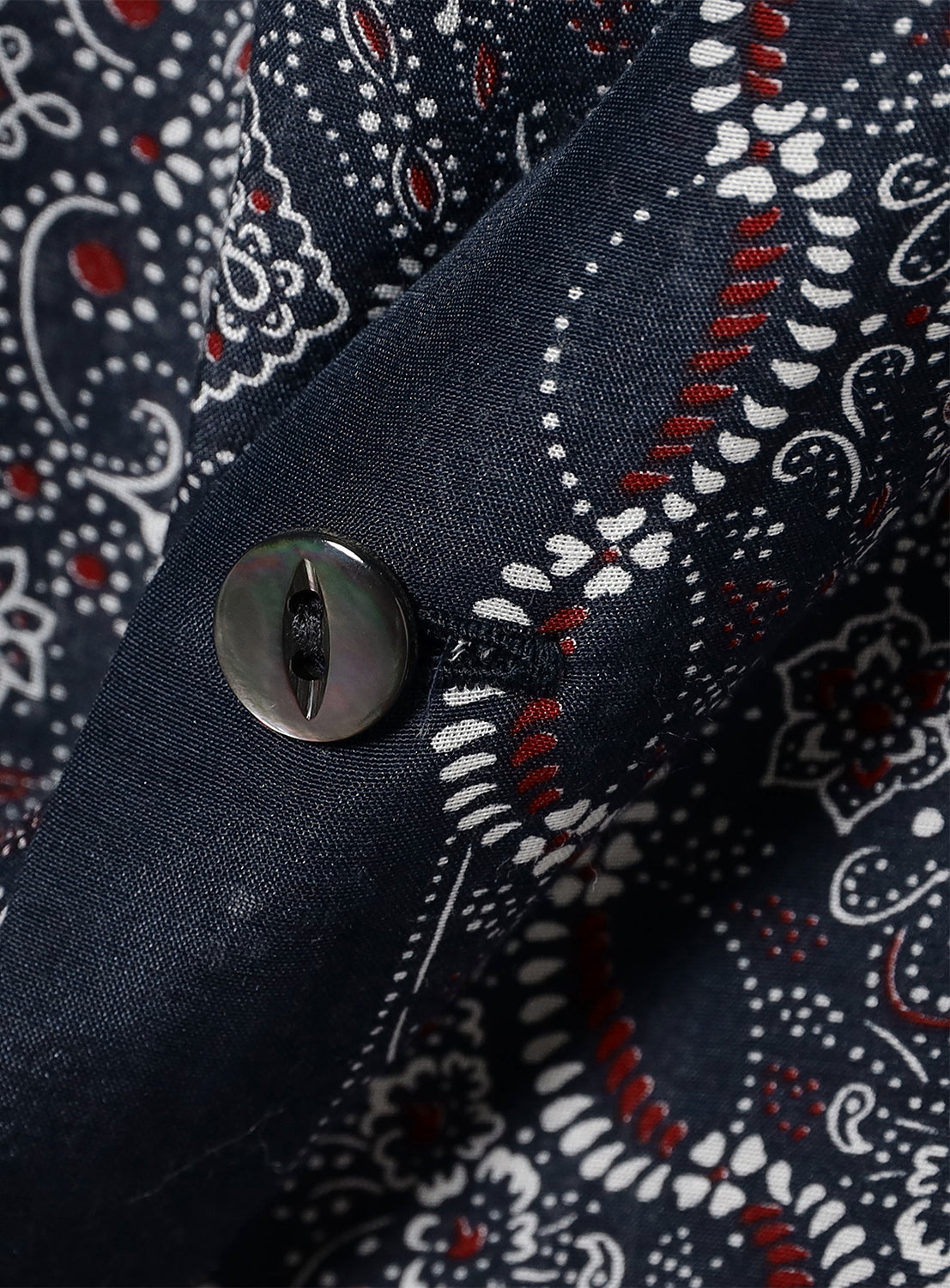 Close-up of navy patterned shirt fabric, detailing the paisley print, red accents, and a mother-of-pearl button.