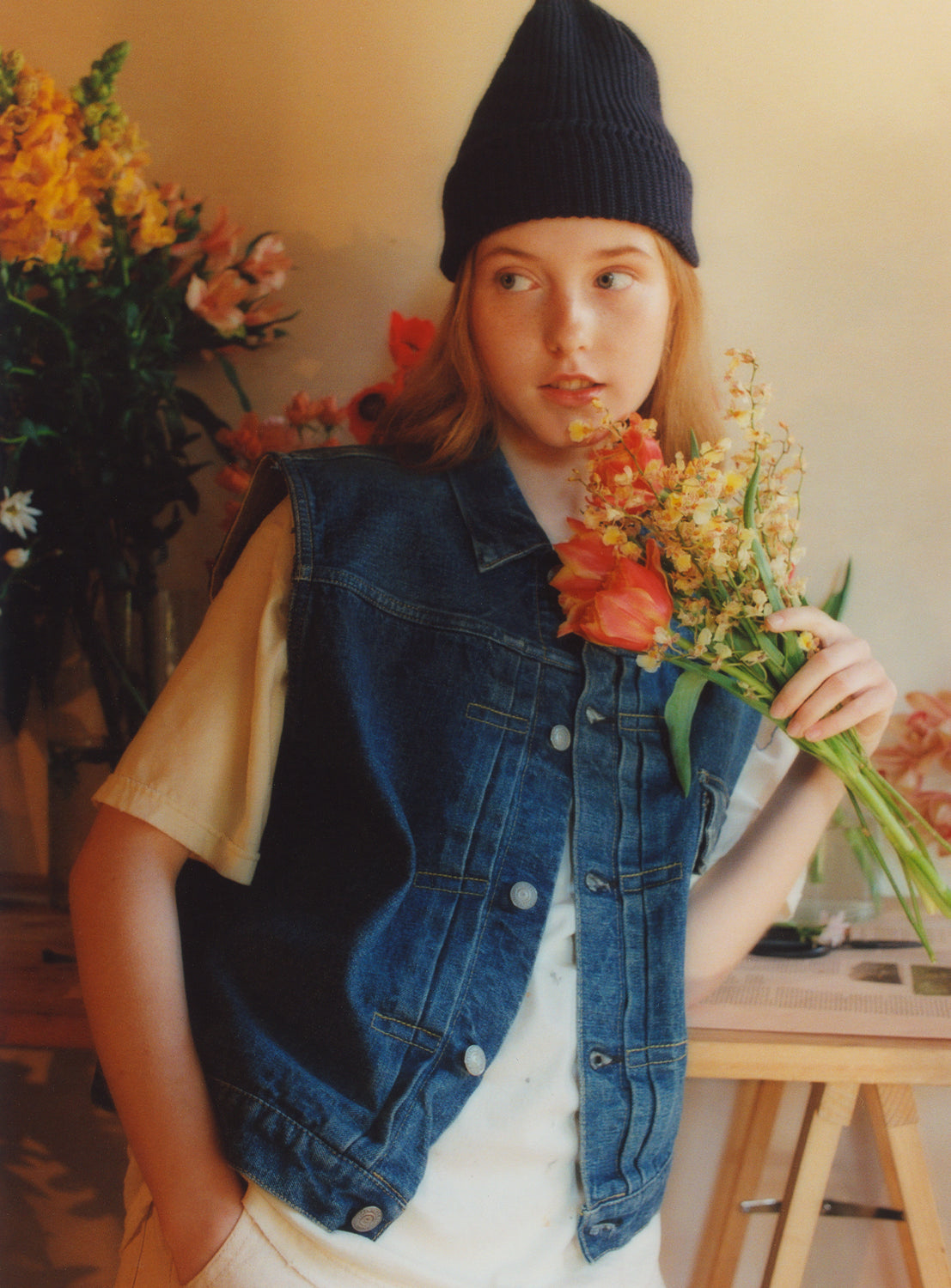 A model wearing a blue denim sleeveless vest, button-up front, and a collar with a white t-shirt underneath.