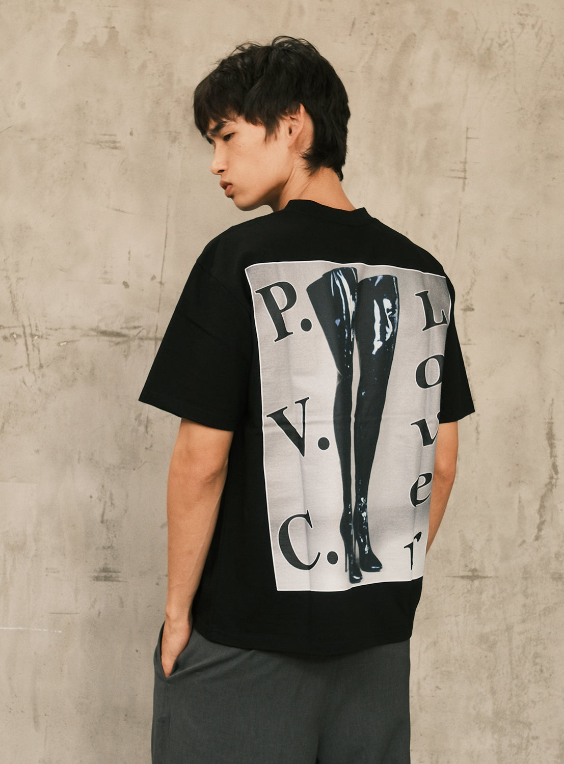 A person wearing the "Sampling For Pleasure Tee" from BIAS DOGS, featuring a black and white graphic of thigh-high boots and the phrase "P.V.C. Lover" on the back, stands against a neutral textured wall.