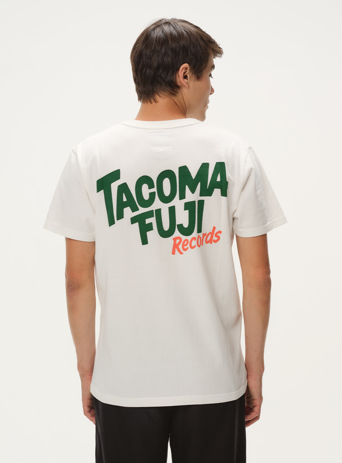 Model wearing a white t-shirt with green and orange "TACOMA FUJI Records" graphic, black shorts, back view.