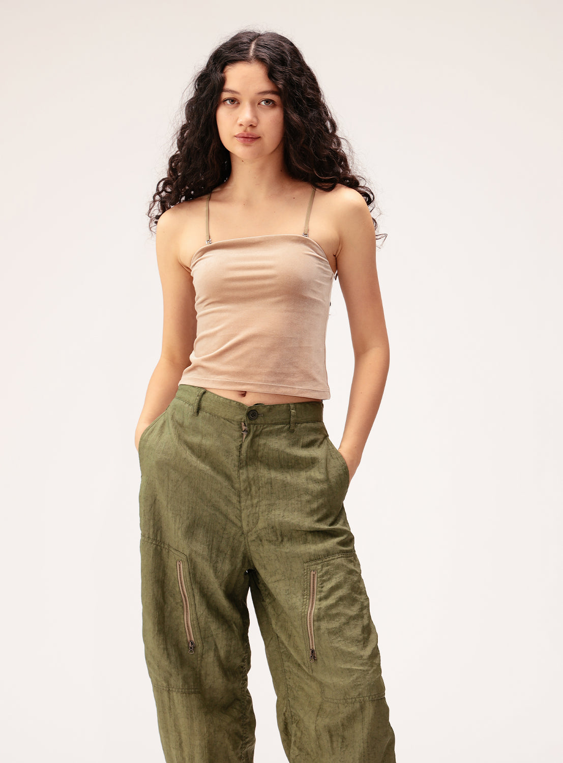 A woman with long, curly dark hair stands against a plain light background with her hands in her pockets, wearing the Ray BEAMS 2-Way Convertible Velour Camisole in beige—featuring spaghetti straps—paired with loose-fitting olive green pants with front zippers.