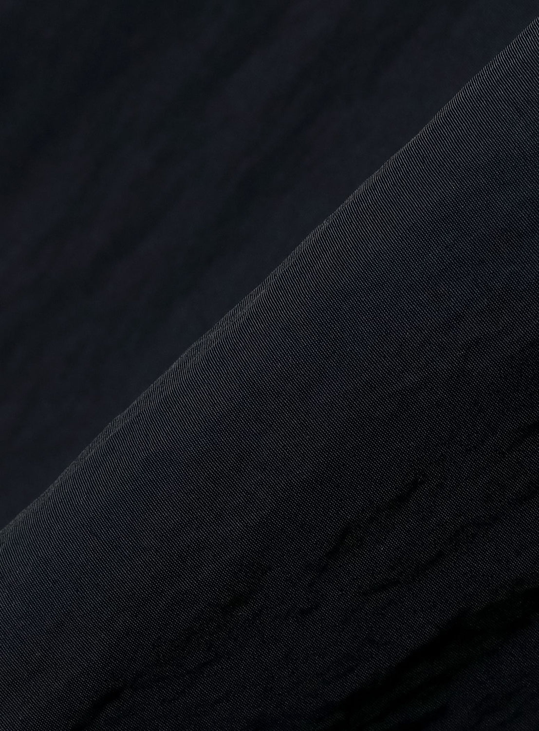 Close-up of the BEAMS T Tyrrell Winston - Anorak Jacket’s dark, textured fabric with diagonal ribbing. The light highlights the fine lines and intricate pattern across the material, revealing subtle shifts in shade from deep black to dark gray—evoking the timeless aesthetic of vintage training wear from this exclusive collaboration.