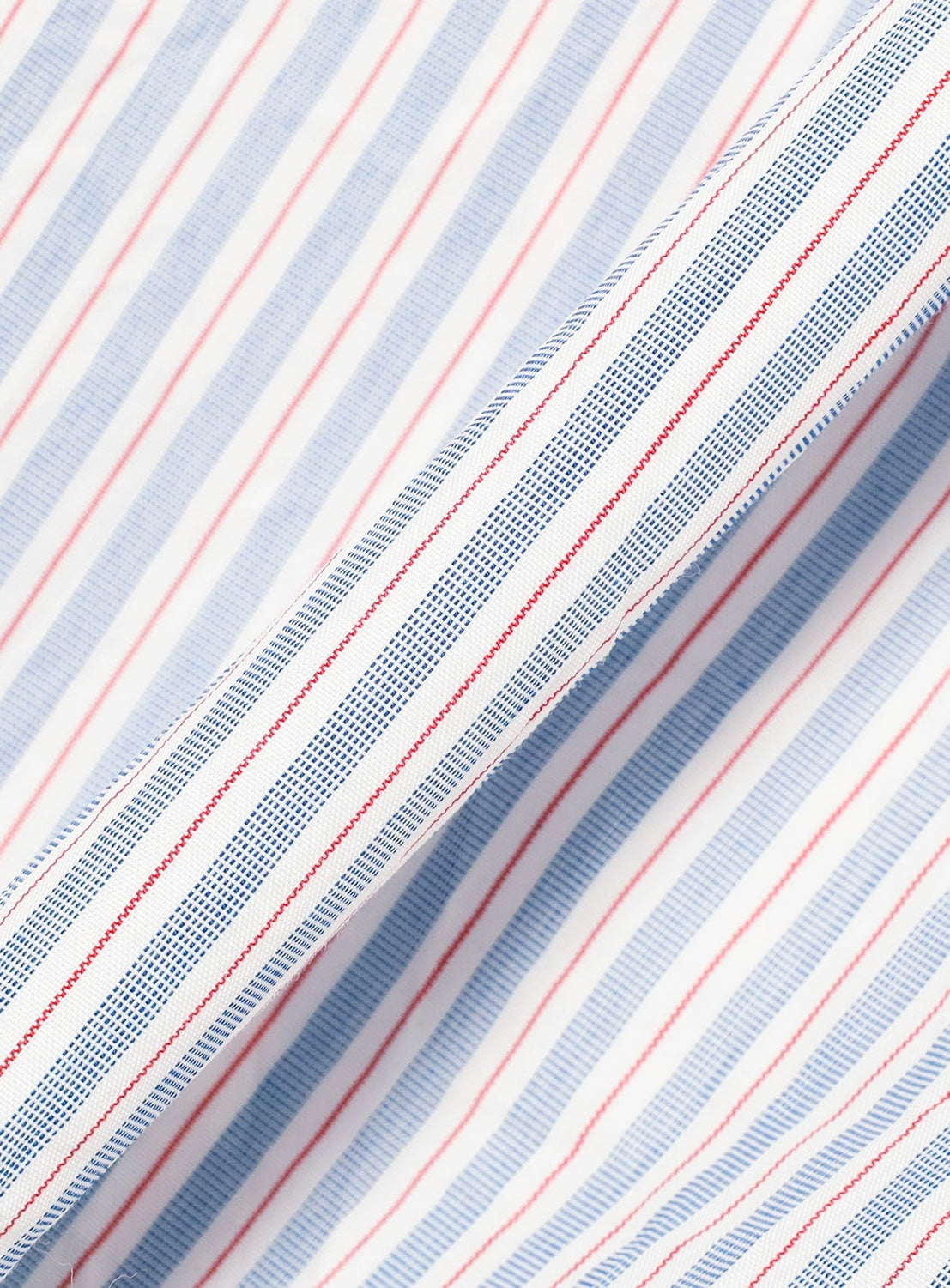 Close-up of the BEAMS BOY Crinkle Stripe Short-Sleeve Dress fabric, featuring diagonal blue, red, and white stripes in varying thicknesses and spacing for a dynamic look. The fabric’s gentle folds highlight its crinkled texture and fluid movement—perfect for a relaxed fit shirt dress.