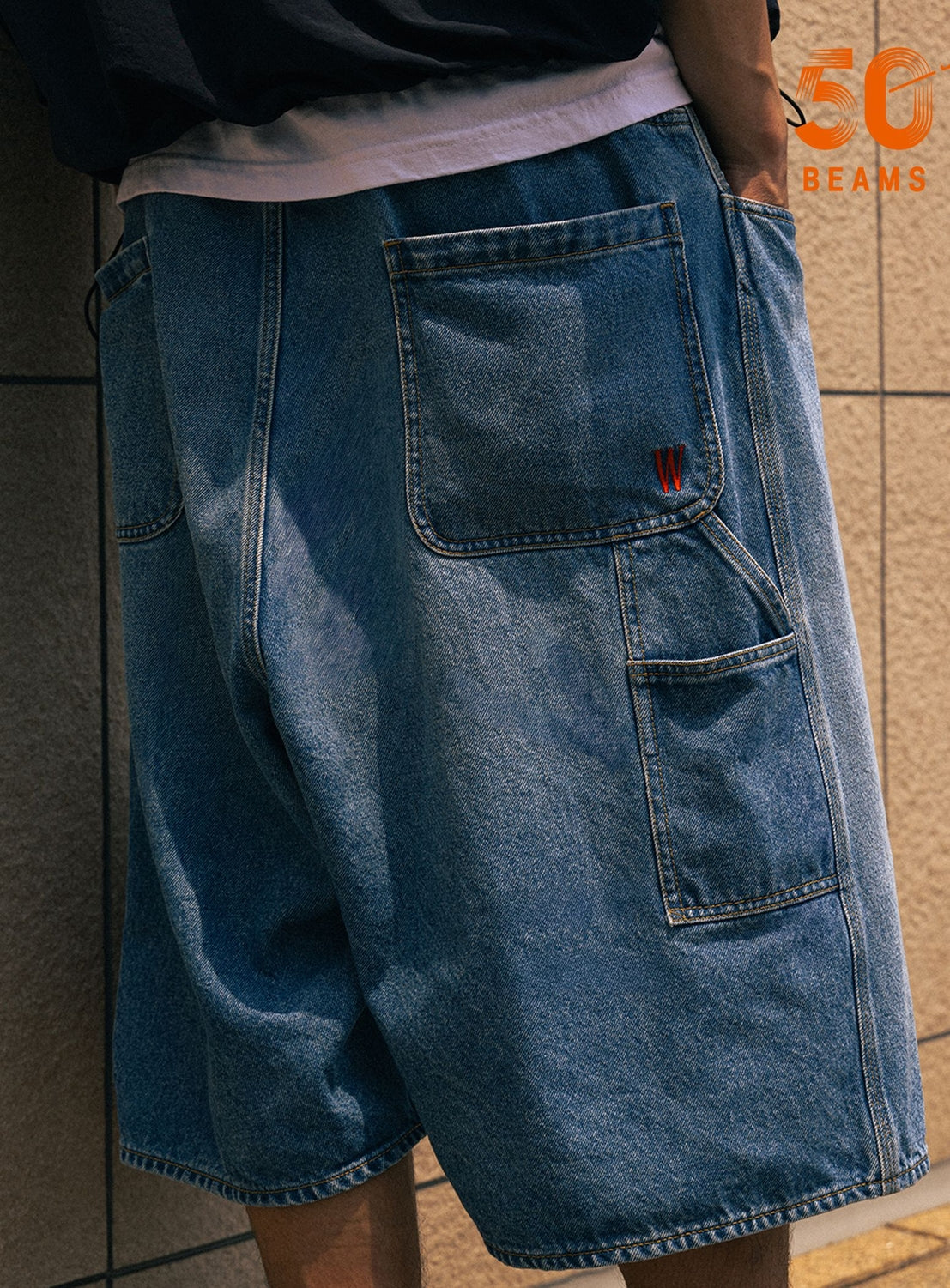 A person wearing loose-fitting Tyrrell Winston - Denim Shorts by BEAMS T, designed with large back pockets and small red embroidery on the upper part, stands against a tiled wall. “50 BEAMS” appears in orange in the top right corner, referencing the BEAMS T x Tyrrell Winston collaboration.