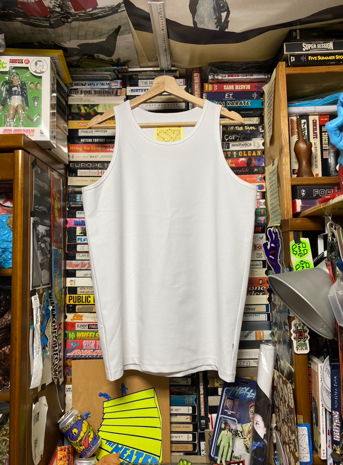 A white tanktop hanging on a hanger with a yellow tag, displayed in front of a shelf with various items.