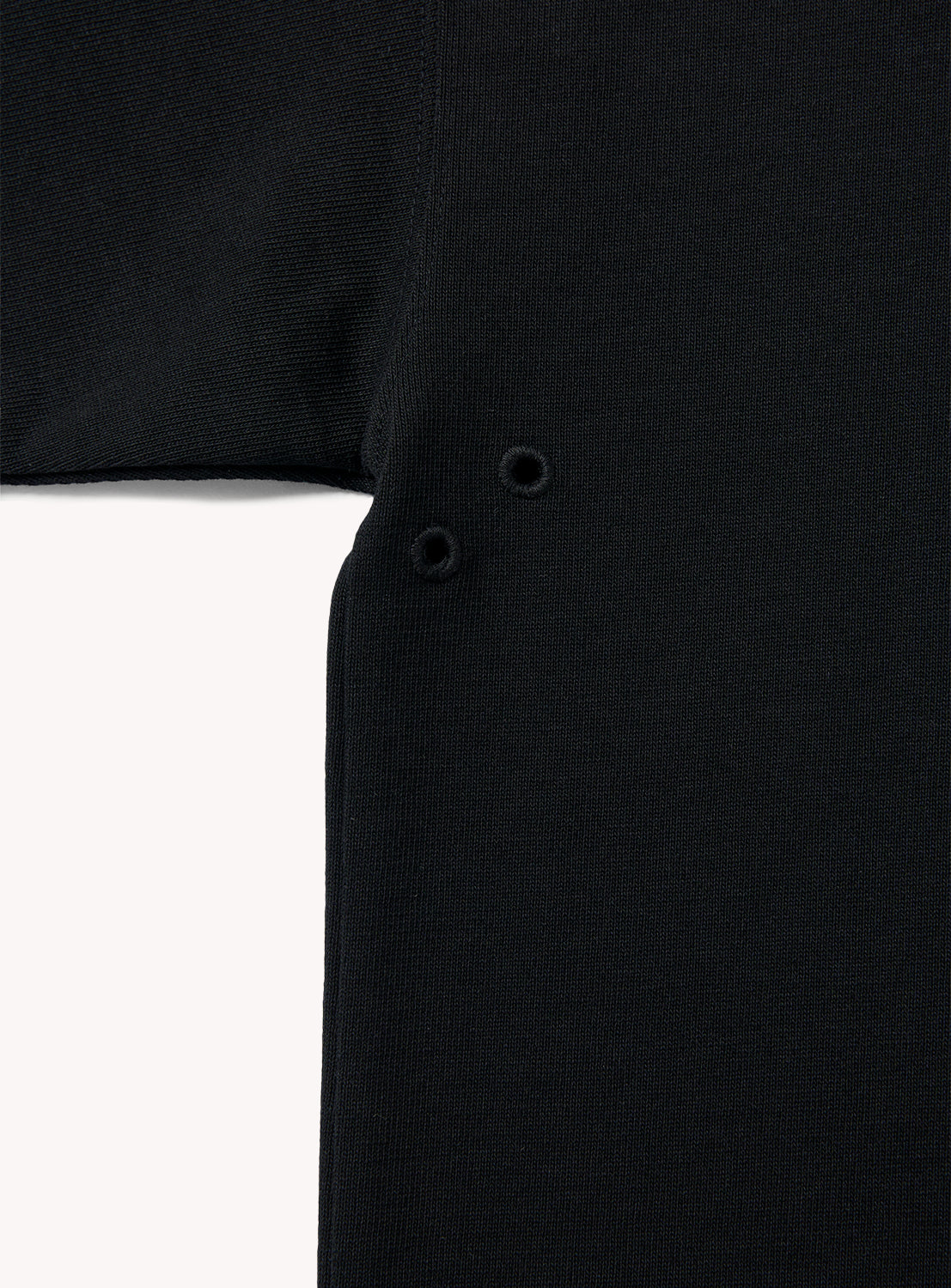 Close-up of the BEAMS "the A - Suvin Turi T-Shirt" in black, featuring two small eyelet holes near the armpit for ventilation. The fabric has a textured knit appearance.