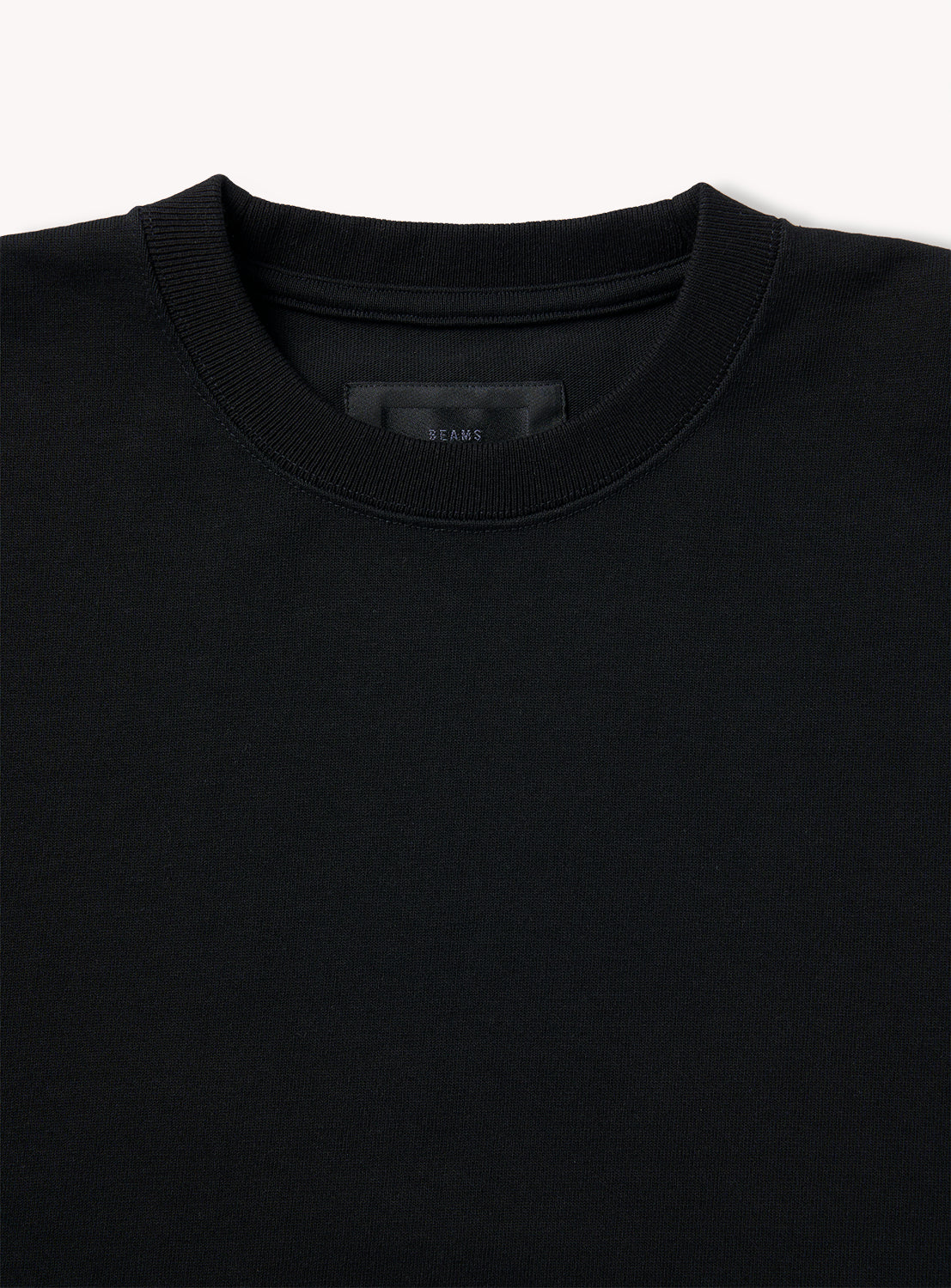 A close-up of the BEAMS Suvin Turi T-Shirt in black, laid flat to show its crew neck collar and upper chest against a light backdrop. The label inside is visible but unreadable, reflecting classic style from the A.