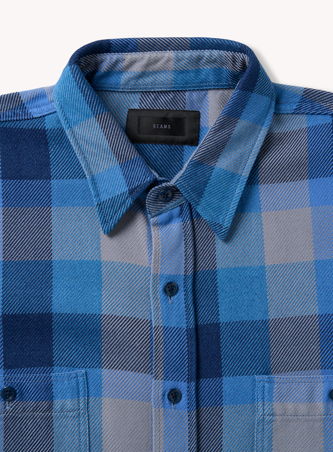 The Indigo Check Shirt by BEAMS features a blue and gray plaid flannel design with a relaxed fit. It is styled with a classic collar, black buttons, and two front chest pockets. The BEAMS label is displayed on the inside of the collar, combining both comfort and style.