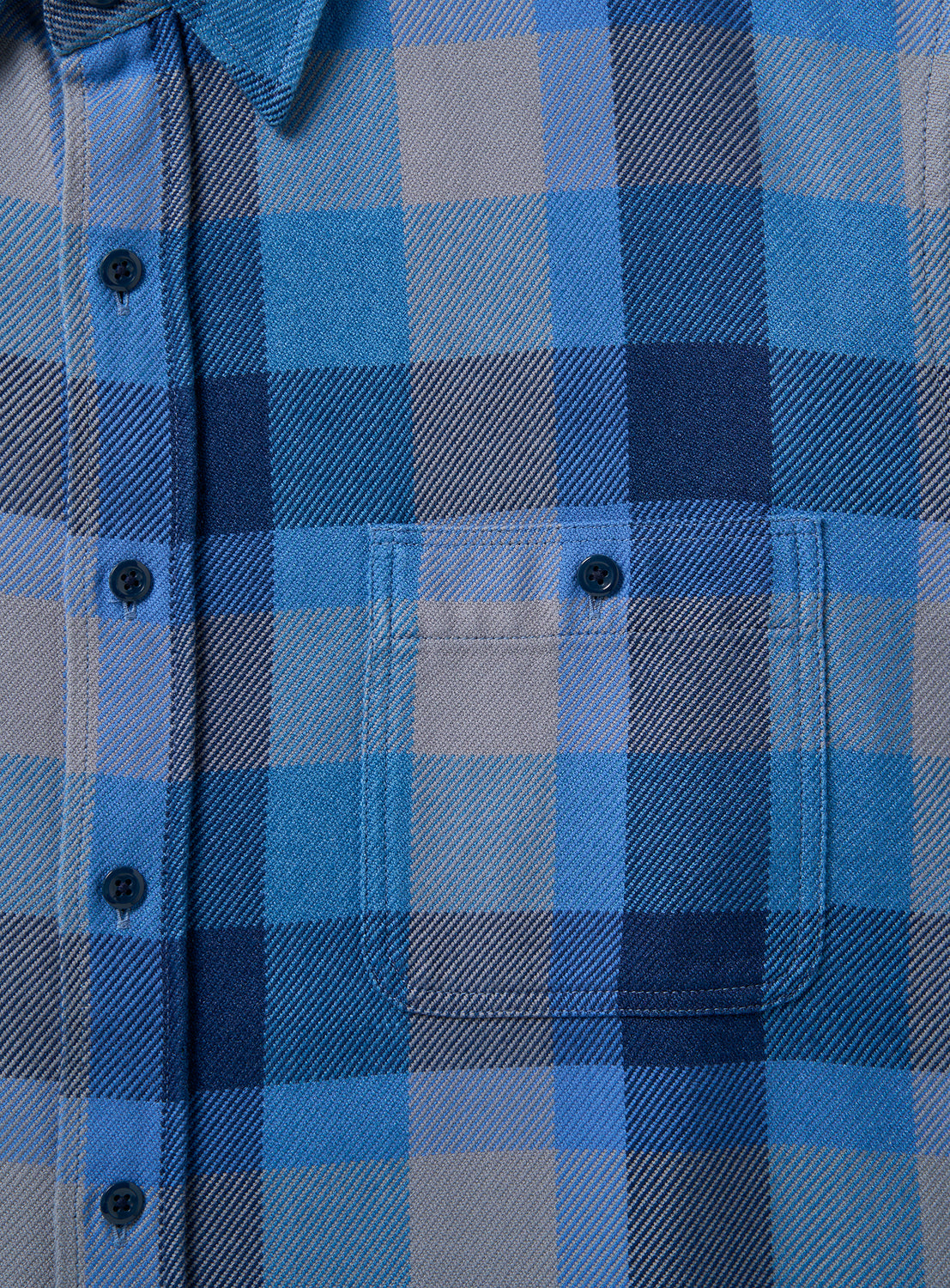 Close-up of the BEAMS Indigo Check Shirt, highlighting its relaxed fit with black buttons and a single chest pocket. The soft fabric displays intersecting lines in various shades of blue and gray, forming a timeless plaid pattern.