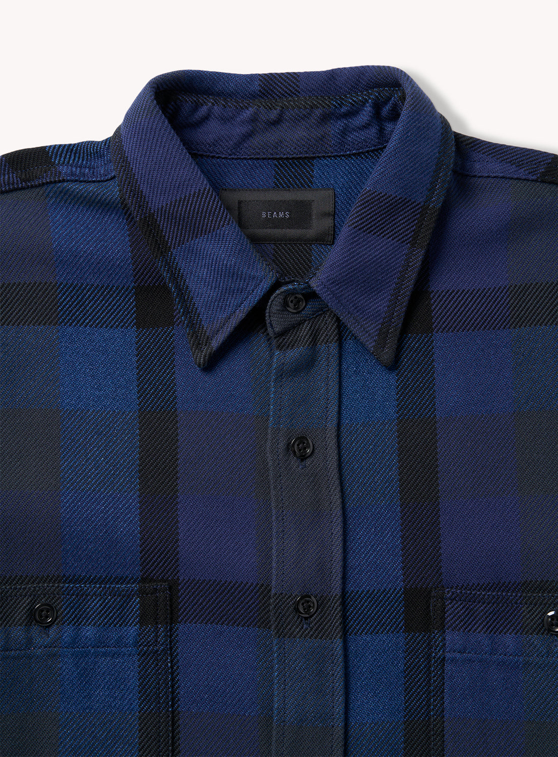 A close-up of the BEAMS Indigo Check Shirt showcases its blue and black plaid pattern, collared neckline, and dual chest pockets. This relaxed fit button-up is neatly arranged and finished with black buttons.