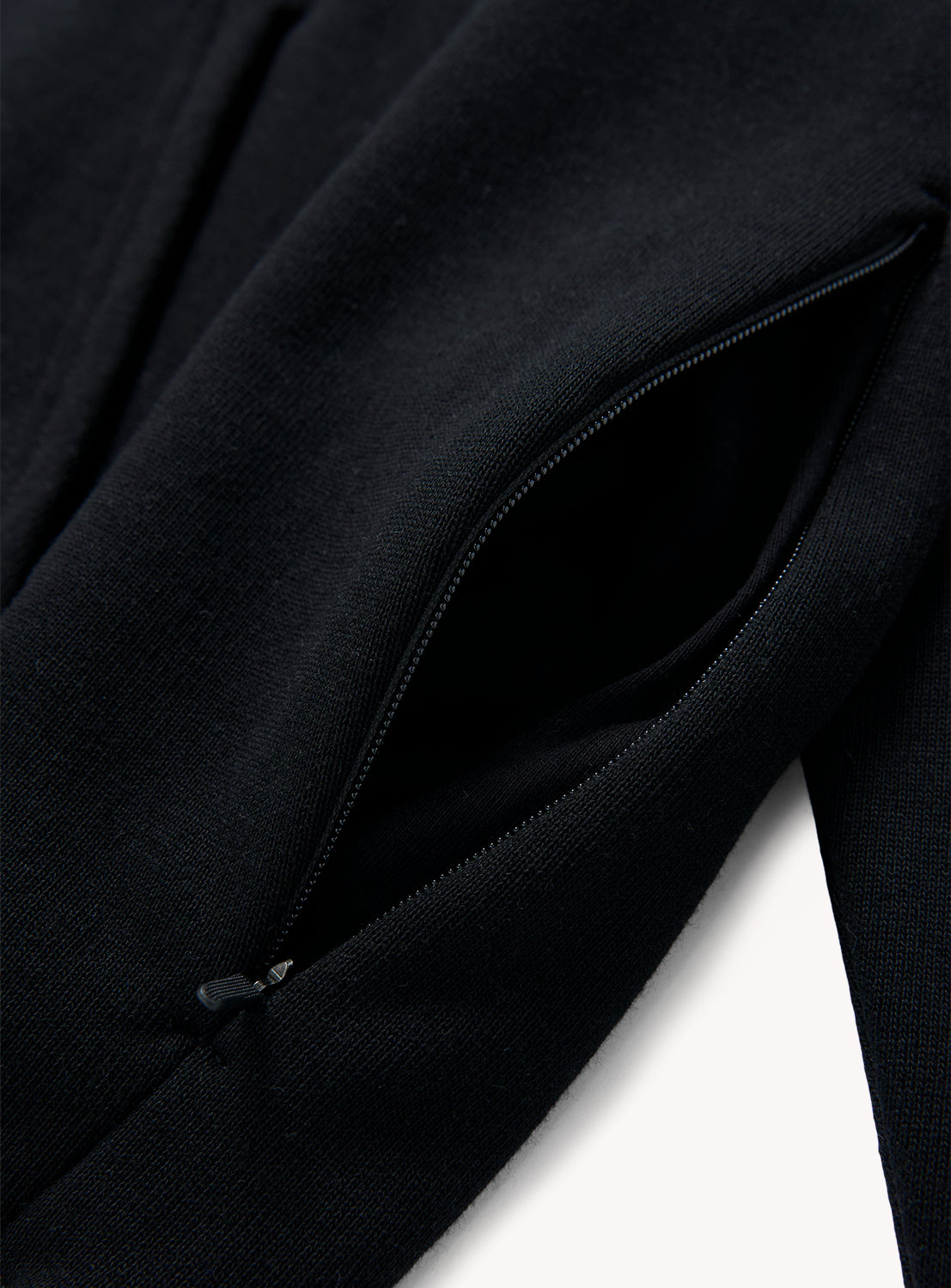 Close-up of the BEAMS Suvin Turi Hoodie, highlighting its minimalist design with a partially unzipped pocket that reveals the zipper teeth and showcases the texture of its premium cotton fabric. The dark garment stands out sharply against the clean white background.