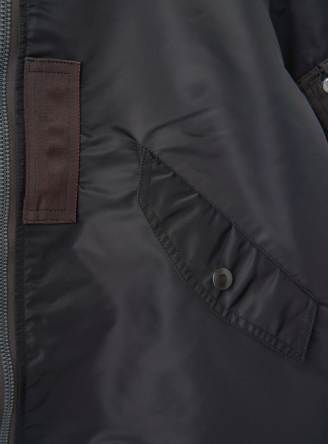 Close-up of a black, shiny William Gibson Collection L-2B jacket by BUZZ RICKSON’s, Made in Japan, featuring a buttoned flap pocket and a brown rectangular patch next to a zipper.