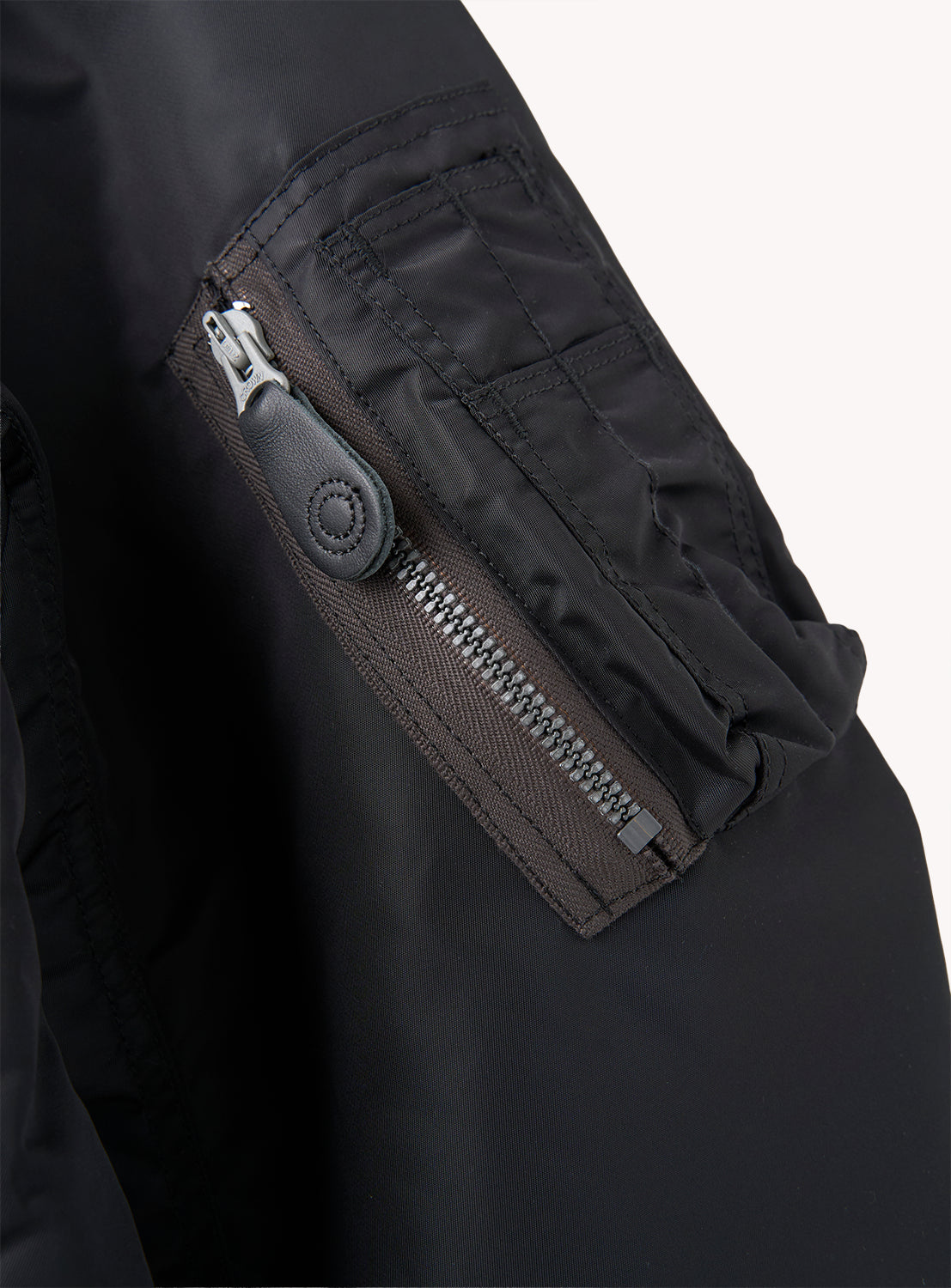 Close-up of a black William Gibson Collection L-2B jacket sleeve by BUZZ RICKSON’s, highlighting a vertical zippered pocket with a leather pull tab and visible stitching details. The jacket features matte-finish fabric and is made in Japan.