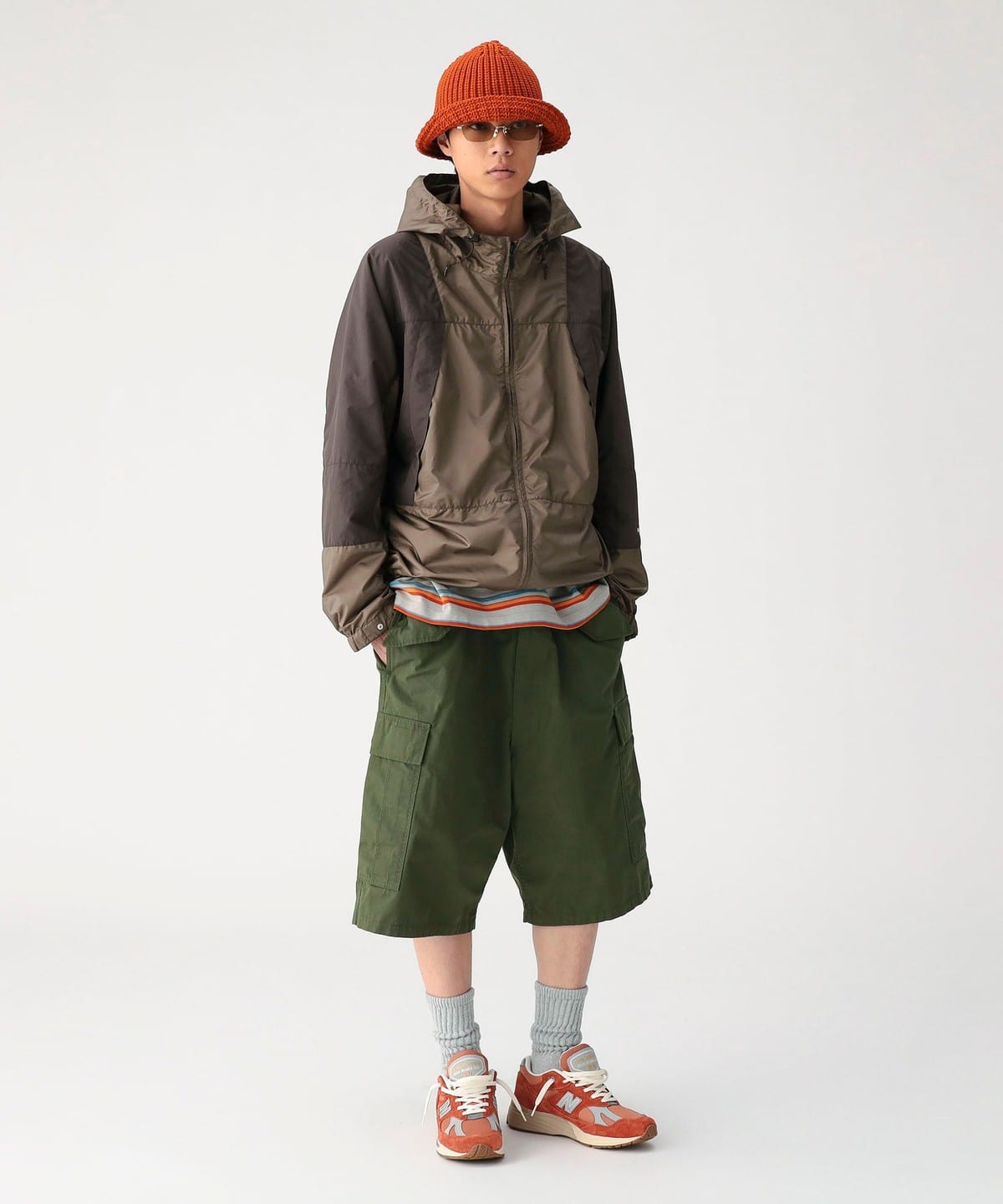 A person wearing an orange knit hat, brown jacket, striped shirt, BEAMS Big 6 Pocket Cargo Shorts in green with a relaxed fit, gray socks, and orange sneakers stands against a plain light background.