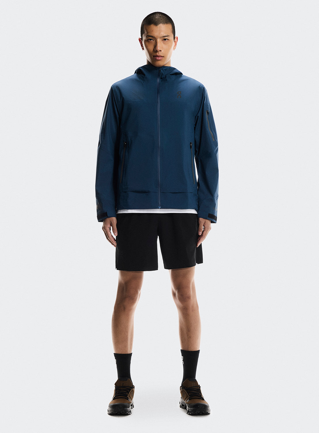Model wearing a blue trek zip-up jacket with multiple zippers. and black shorts. 