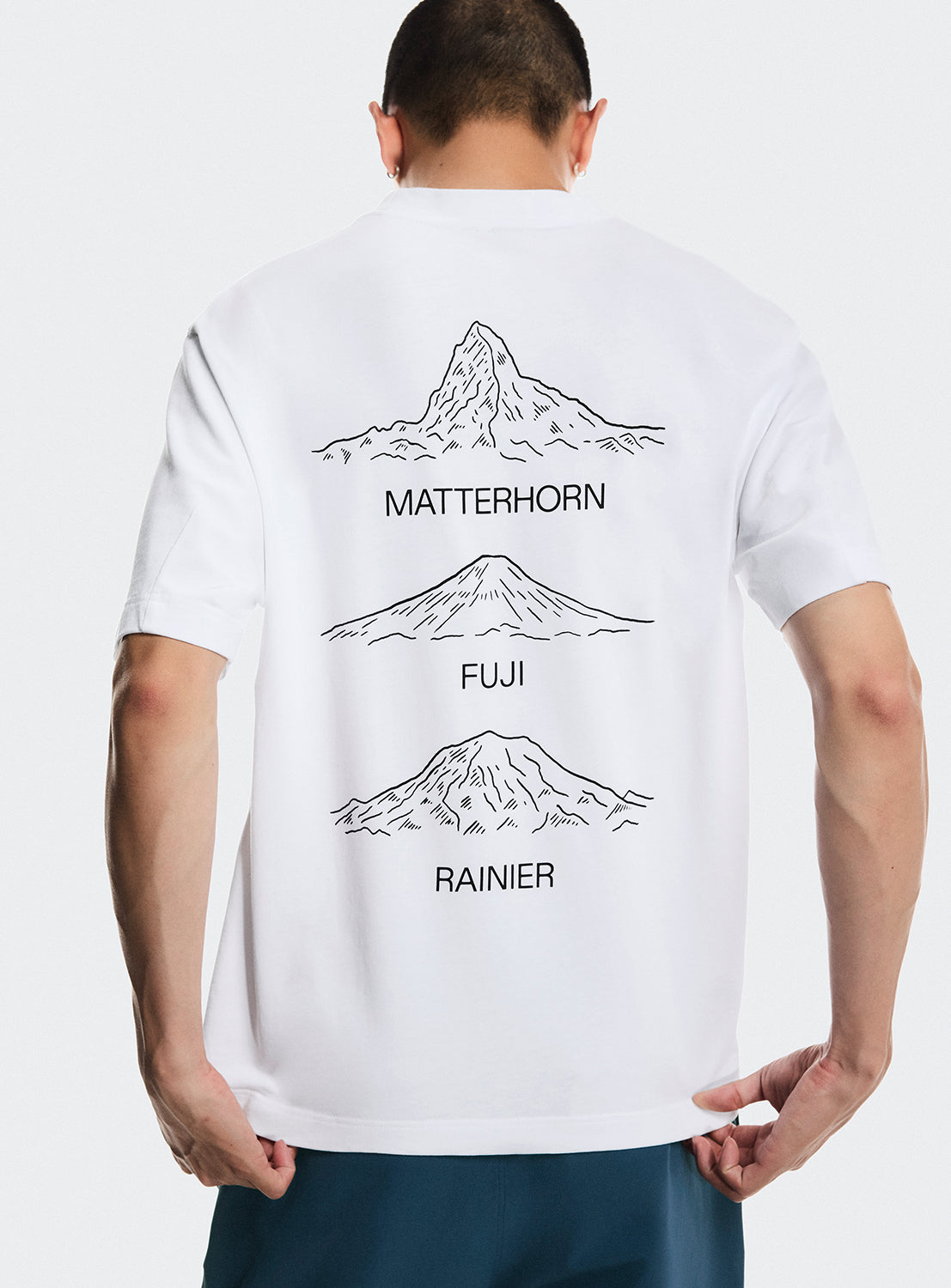 Model wearing a white t-shirt with Matterhorn mountain, Mt. Fuji and Mt. Rainier graphics on the back. 