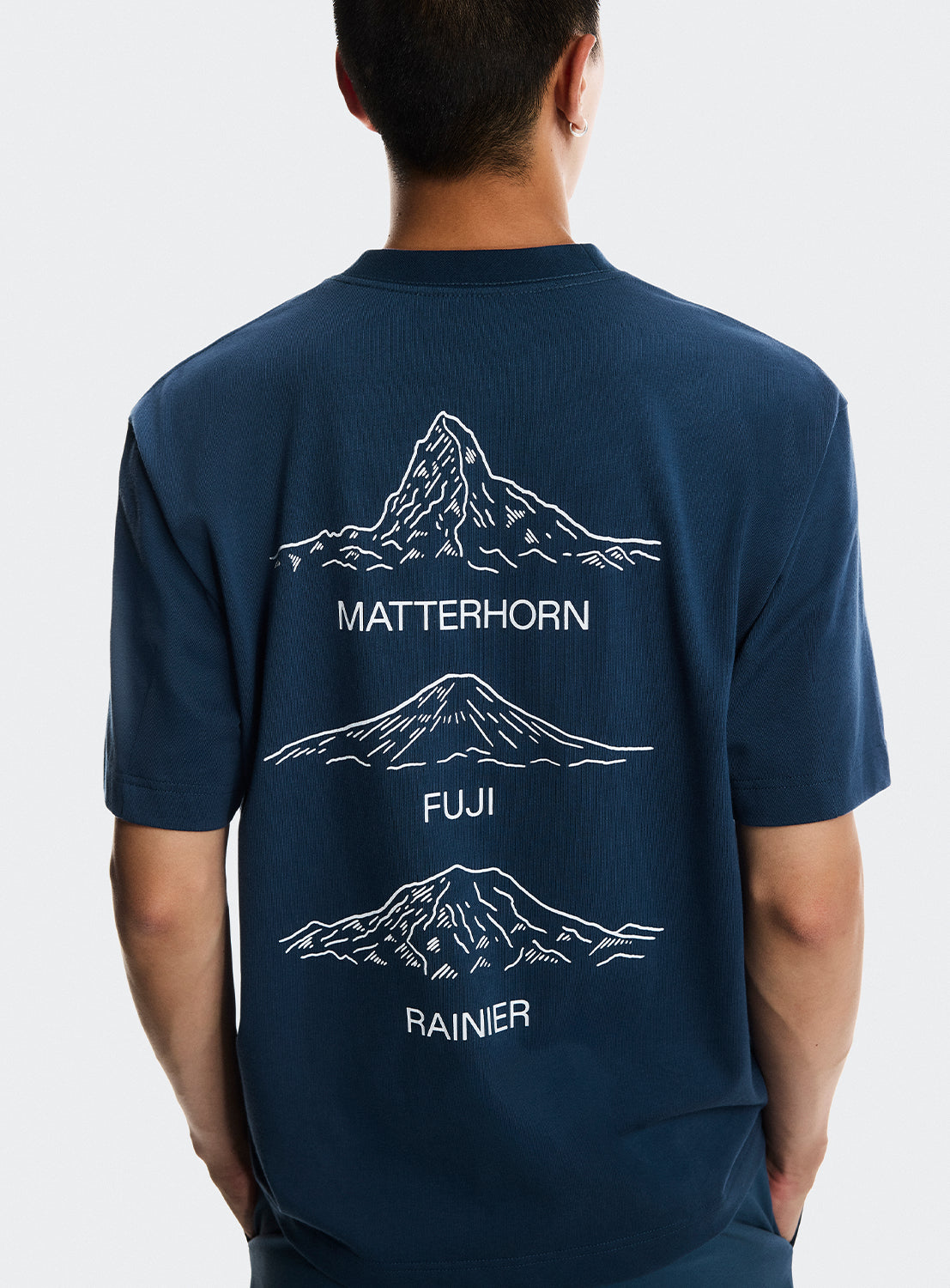 Model wearing navy blue t-shirt with Matterhorn mountain, Mt. Fuji and Mt. Rainier graphics on the back. 