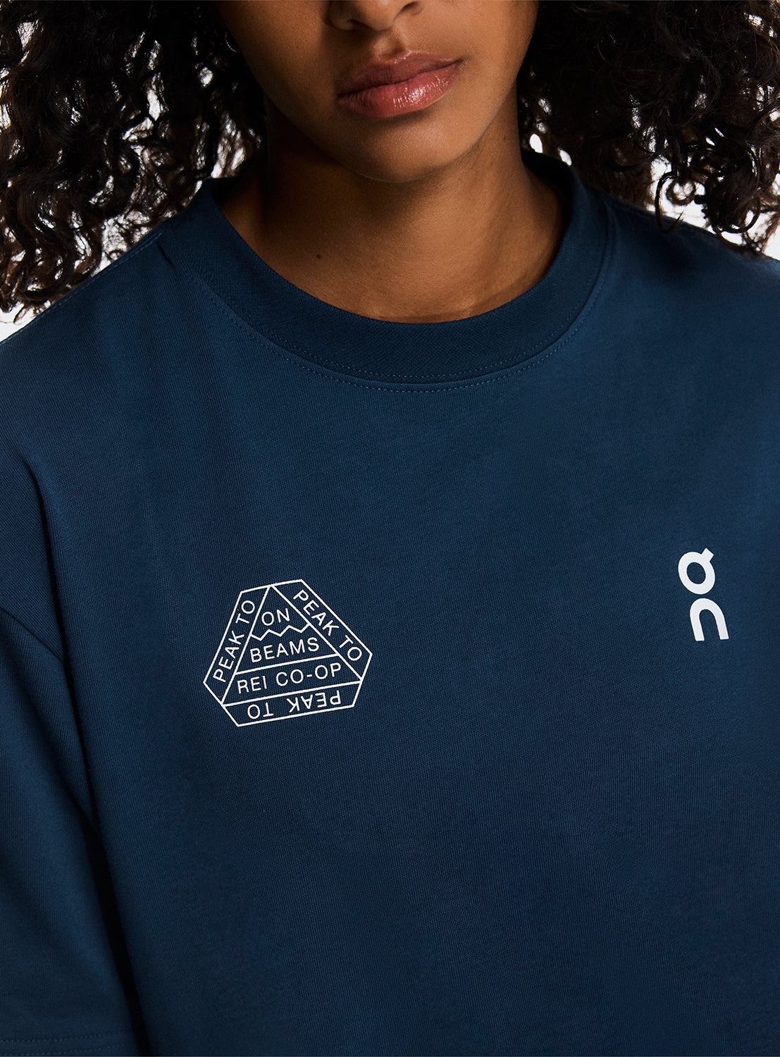 Model wearing navy blue t-shirt with close up of ON Running, BEAMS, and REI CO-OP logos. 