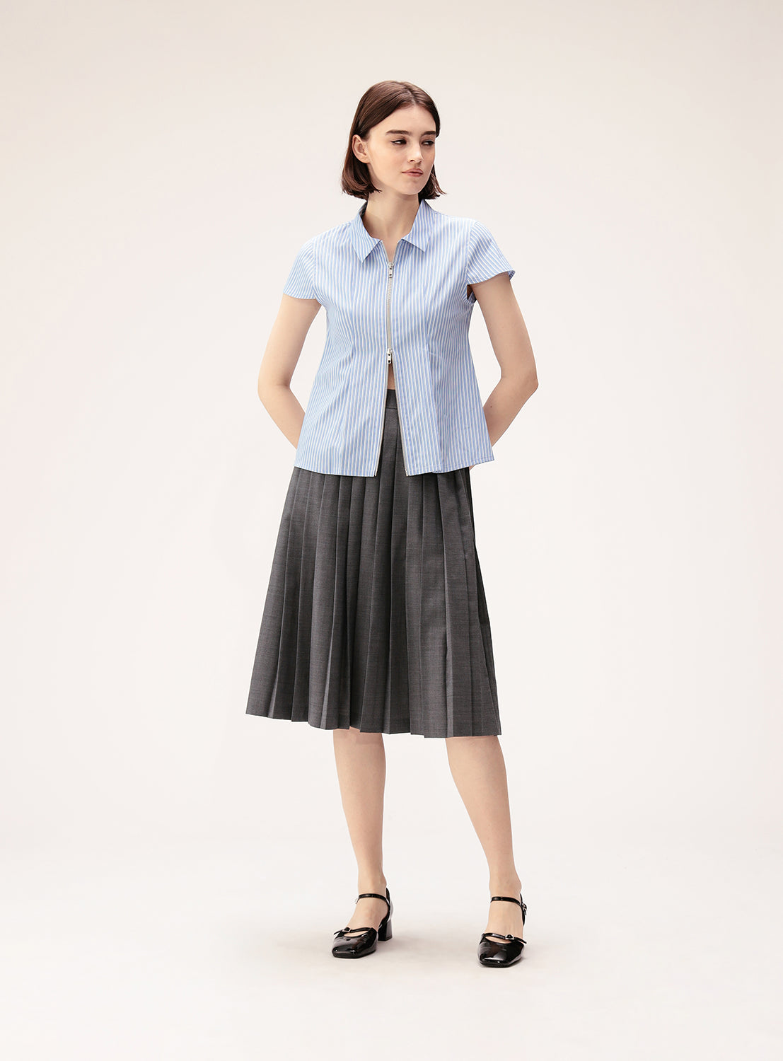 A woman stands in a studio wearing a short-sleeve blue striped shirt, black shoes, and the BEAMS BOY Mixed Pleat Skirt—a gray pleated midi skirt crafted from lightweight fabric—with her hands in her pockets.