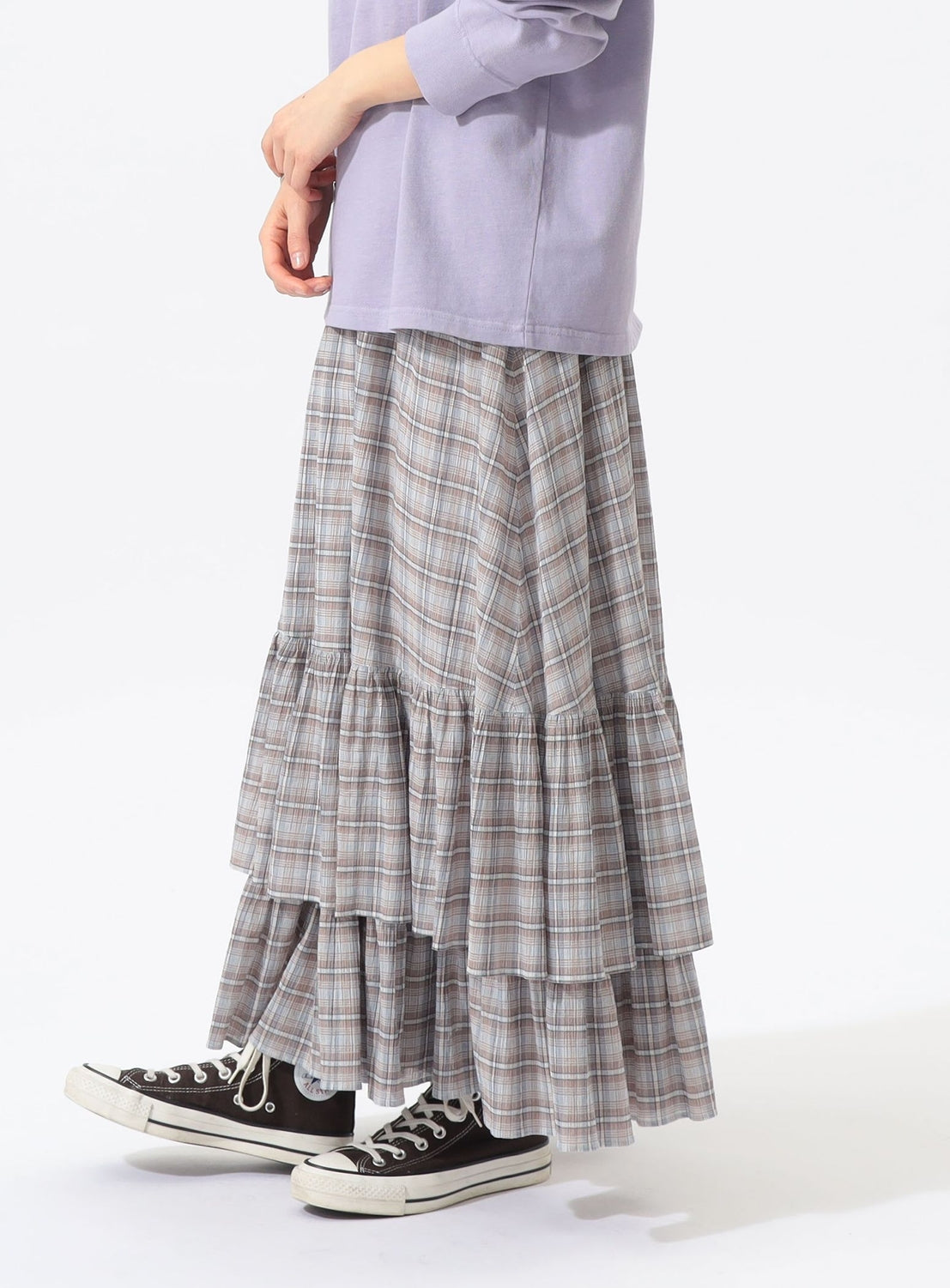 A person is shown from the waist down, standing against a plain white background, wearing a lavender long-sleeve top paired with the BEAMS BOY Check Tiered Skirt in a lightweight gray plaid fabric and black low-top Converse sneakers.