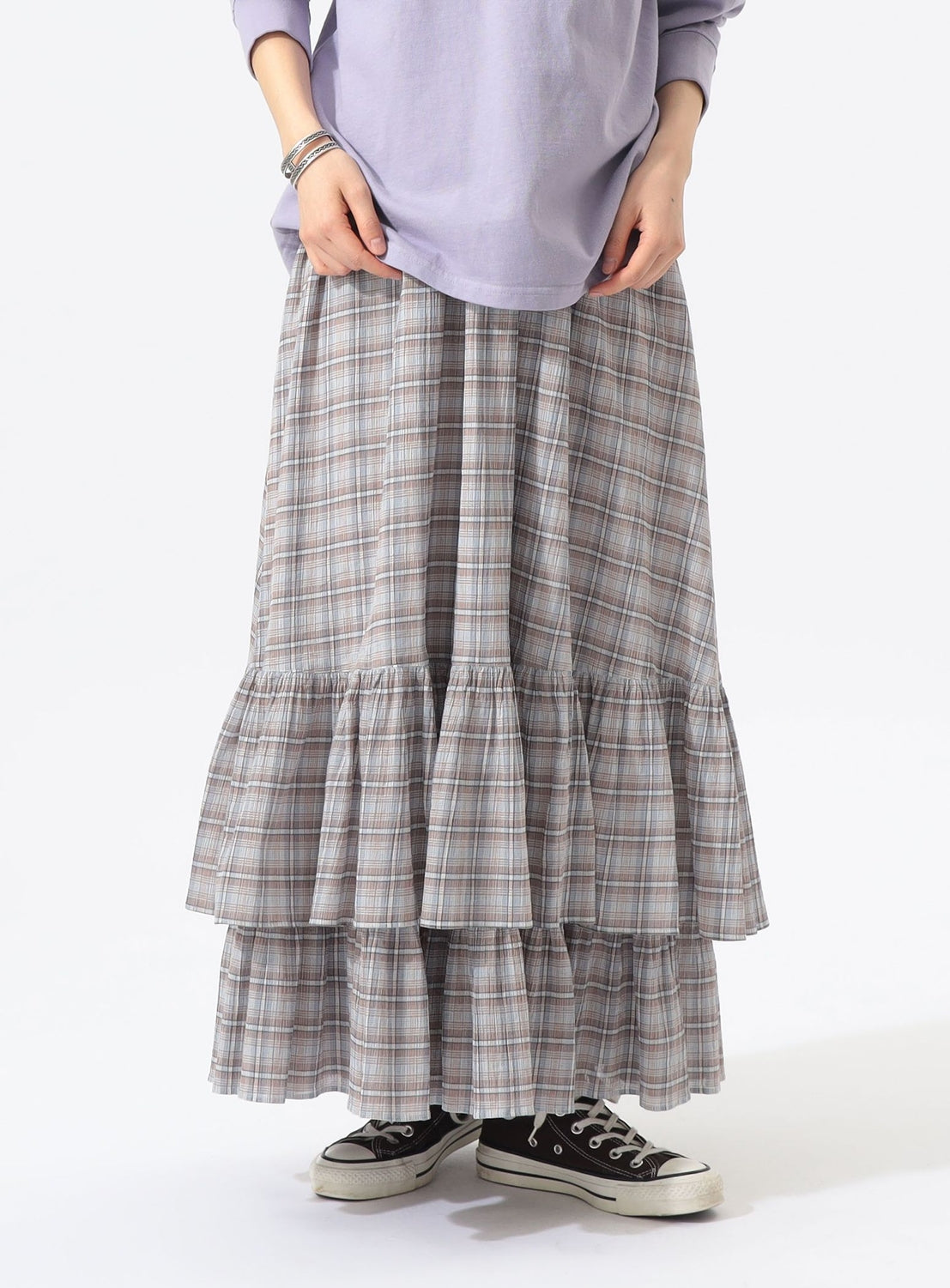 A person is shown from the waist down against a plain background, wearing a light purple long-sleeve top, the BEAMS BOY Check Tiered Skirt in a gray check pattern made of lightweight fabric, and black-and-white sneakers. Their hands are positioned in front of them.