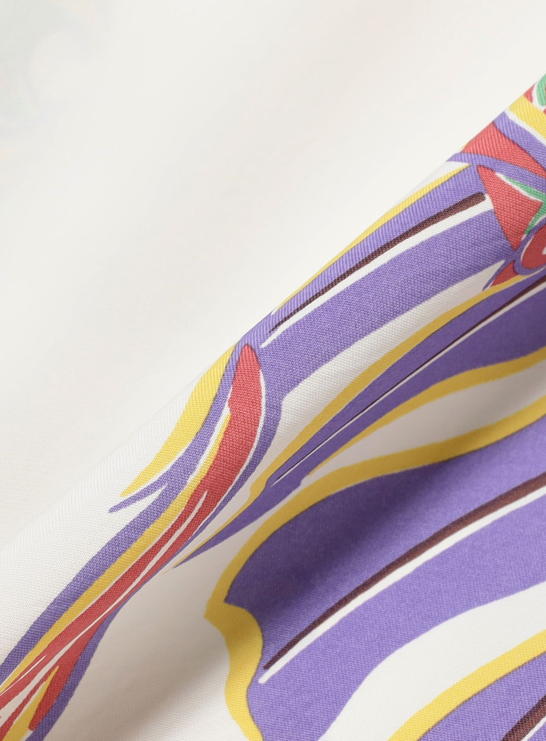A close-up of the [Exclusive] SUN SURF x BEAMS BOY Sailor Shirt fabric shows a colorful abstract pattern with swirling purple, yellow, and red lines on white, inspired by Hawaiian prints. The smooth texture is visible where the fabric is folded.