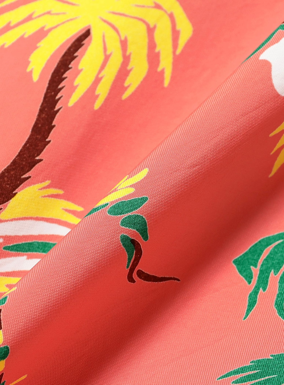 Close-up of the SUN SURF x BEAMS BOY Sailor Shirt’s coral pink fabric. Tropical yellow palm leaves, green fronds, and brown trunks feature on this vintage-inspired material, with visible folds accentuating its texture.