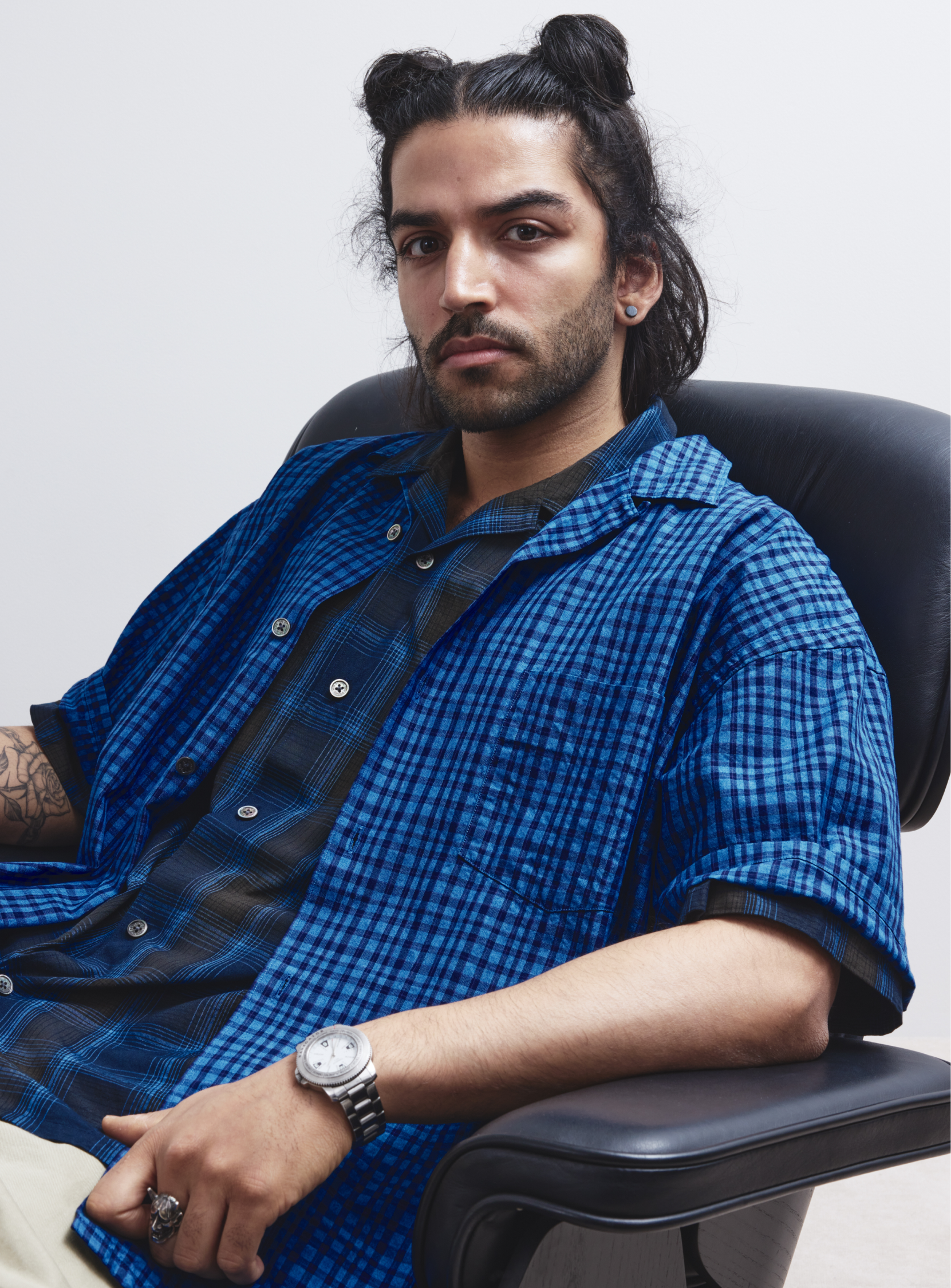 A man with long dark hair in two buns sits in a black chair, wearing the BEAMS [Exclusive] The A - Indigo Check Open Collar Shirt, beige pants, and a watch. He has a serious expression and visible arm tattoos.
