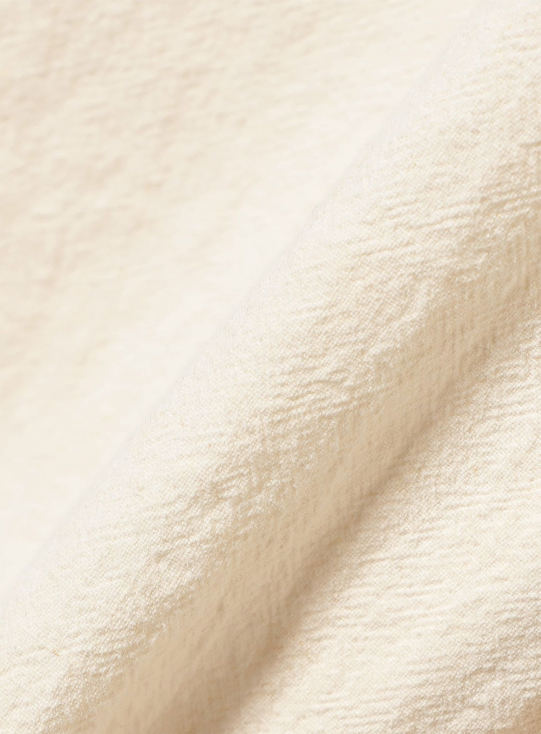 A close-up of the textured, off-white fabric surface from the BEAMS BOY Cotton Linen Trench Coat, featuring soft shadows and diagonal folds that echo the refined structure of classic double-breasted tailoring.