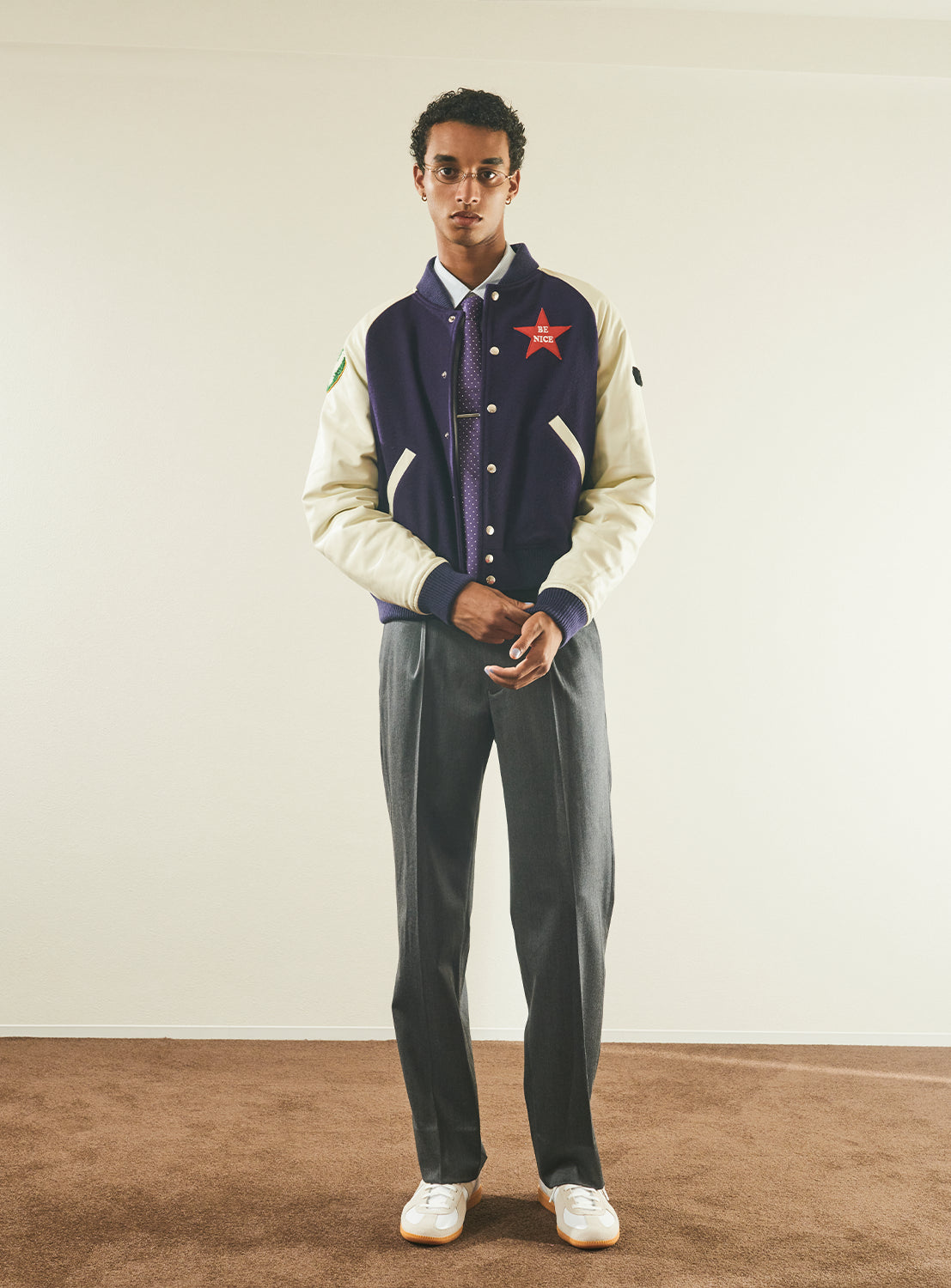 A person stands indoors on brown carpet, wearing the TTT MSW Varsity Patch Jacket in purple and cream, paired with grey trousers, white sneakers, and a tie, looking directly at the camera against a plain light-colored wall.
