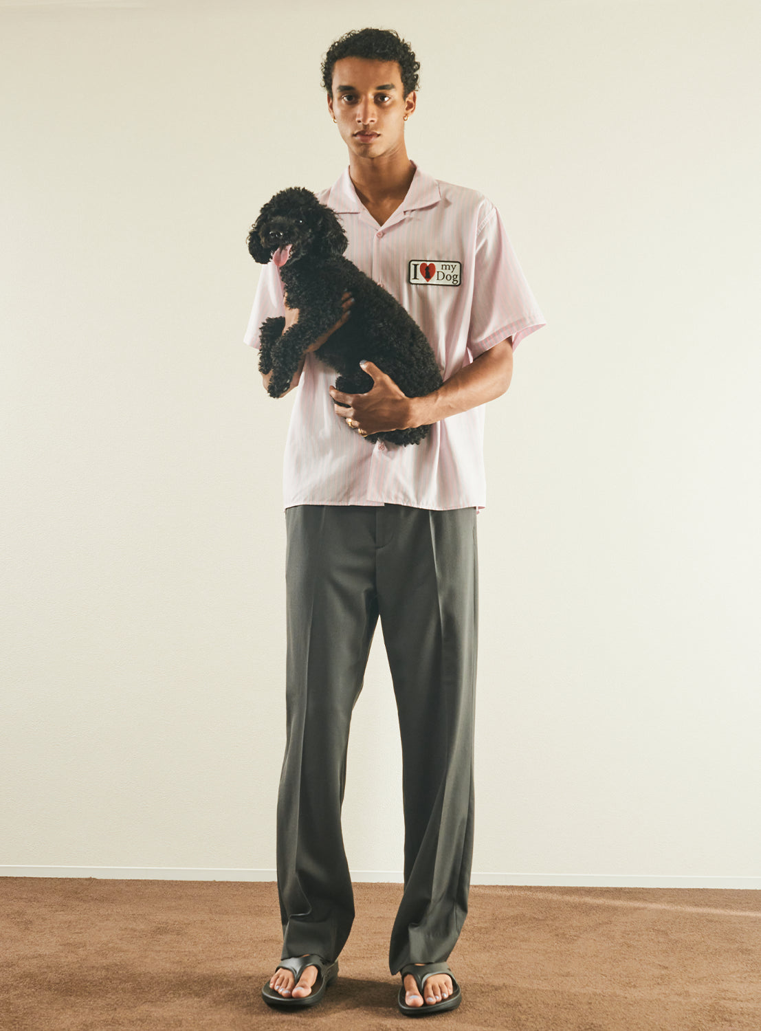 A young man stands indoors holding a small black dog. He wears the TTT MSW “I Love My Dog” Half Sleeve Shirt in light pink, paired with dark gray pants and flip-flops, and is facing the camera with a neutral expression.