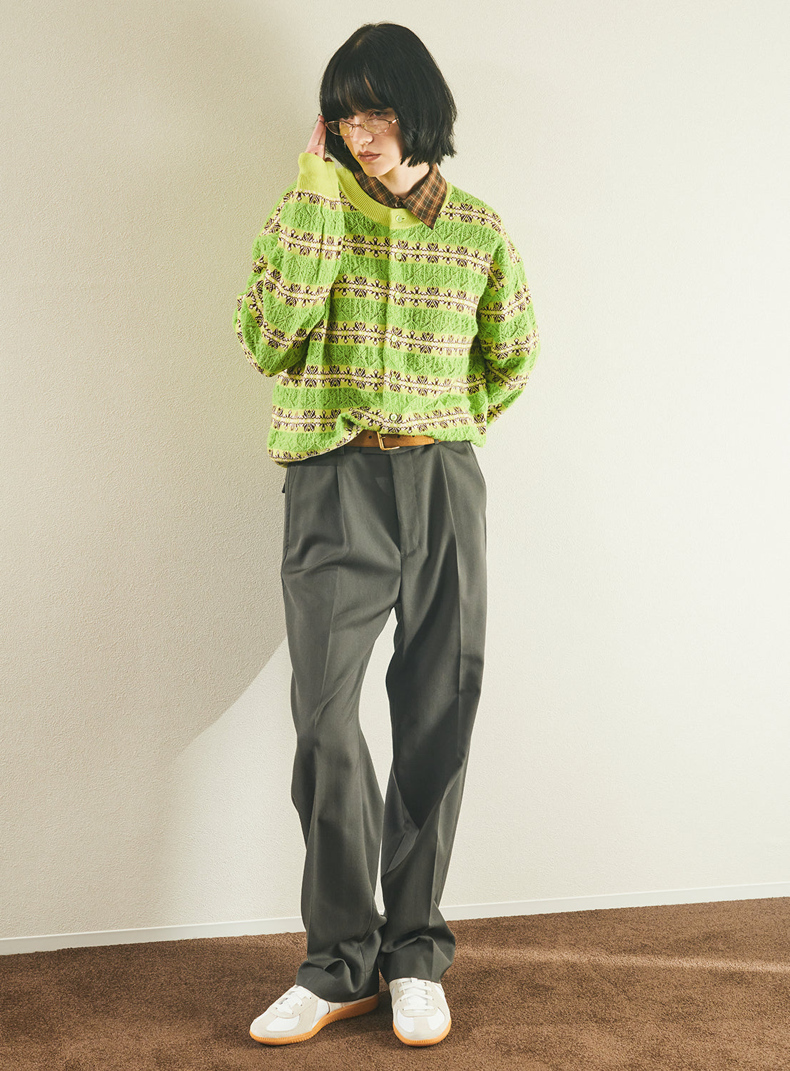 A person with a black bob haircut and glasses is shown standing indoors on a brown carpet, wearing the TTT MSW Crewneck Cardigan in green jacquard, paired with loose gray trousers and white sneakers. They are touching their face with one hand.