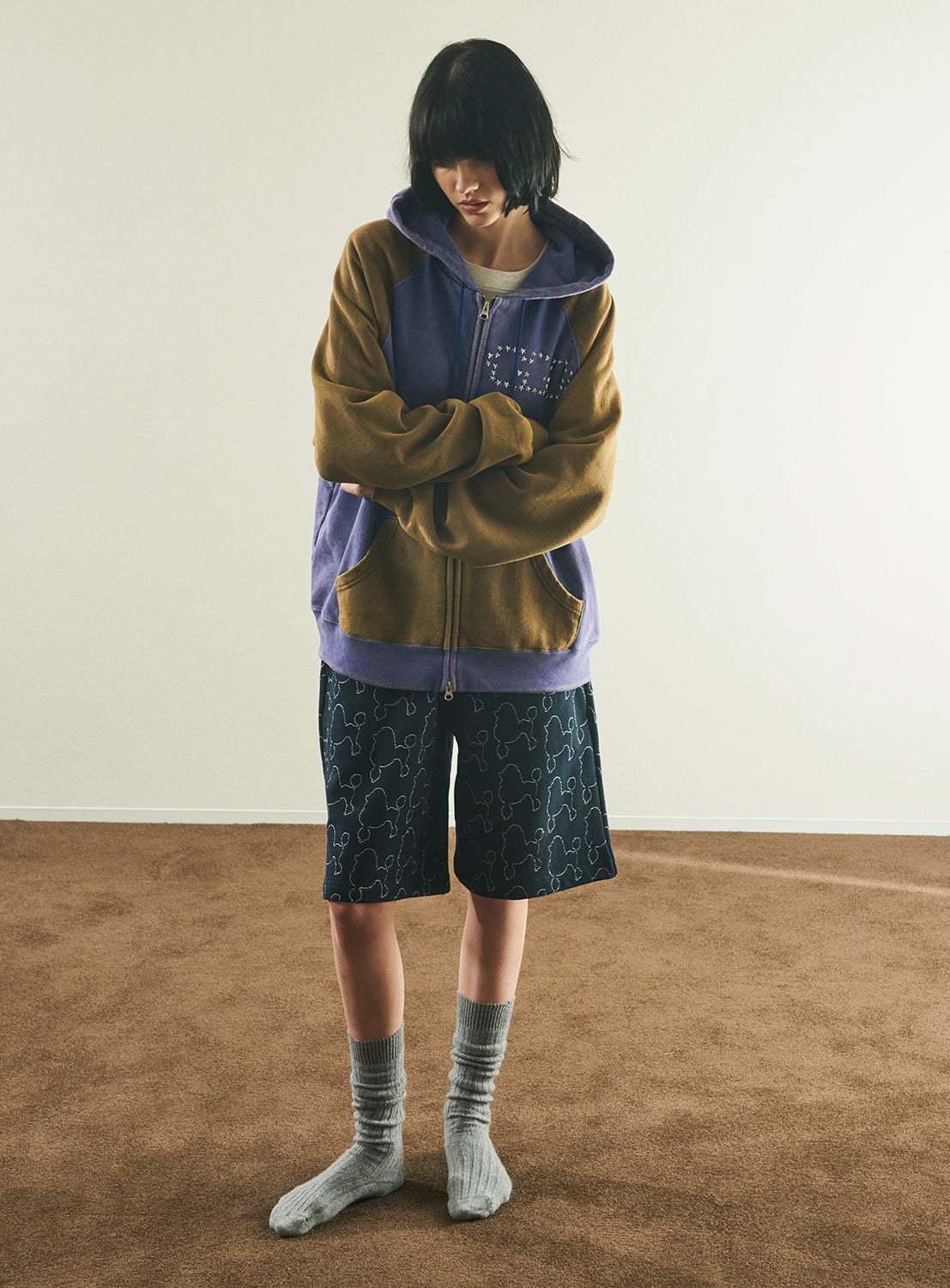 A person with short dark hair stands indoors on a brown carpet, wearing the TTT MSW Fade Damage Zip Up Hoodie—a vintage-inspired piece in shades of purple and brown—paired with black patterned shorts and gray socks. With arms crossed and head slightly bowed, they pose against a plain off-white background.