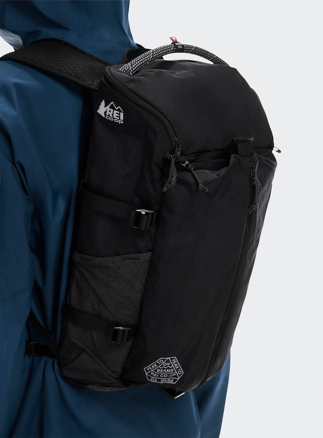 Black backpack with REI and BEAMS logo worn by a person in a blue jacket.